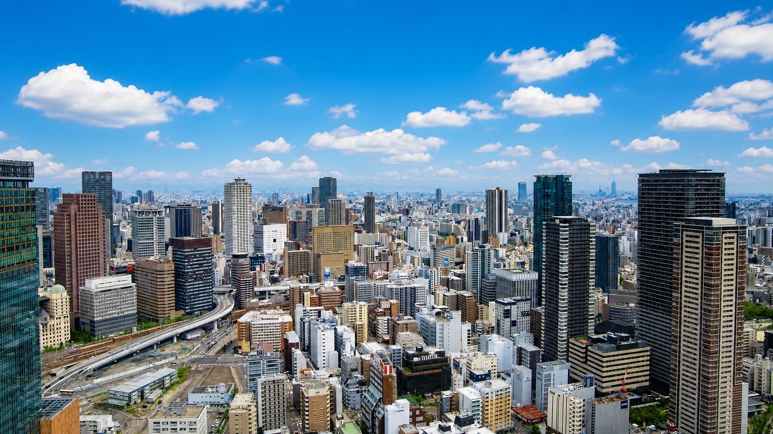 Aerial view over Osaka's dense downtown skyline of high-rise towers and elevated roads under a bright blue sky