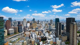 Aerial view over Osaka's dense downtown skyline of high-rise towers and elevated roads under a bright blue sky