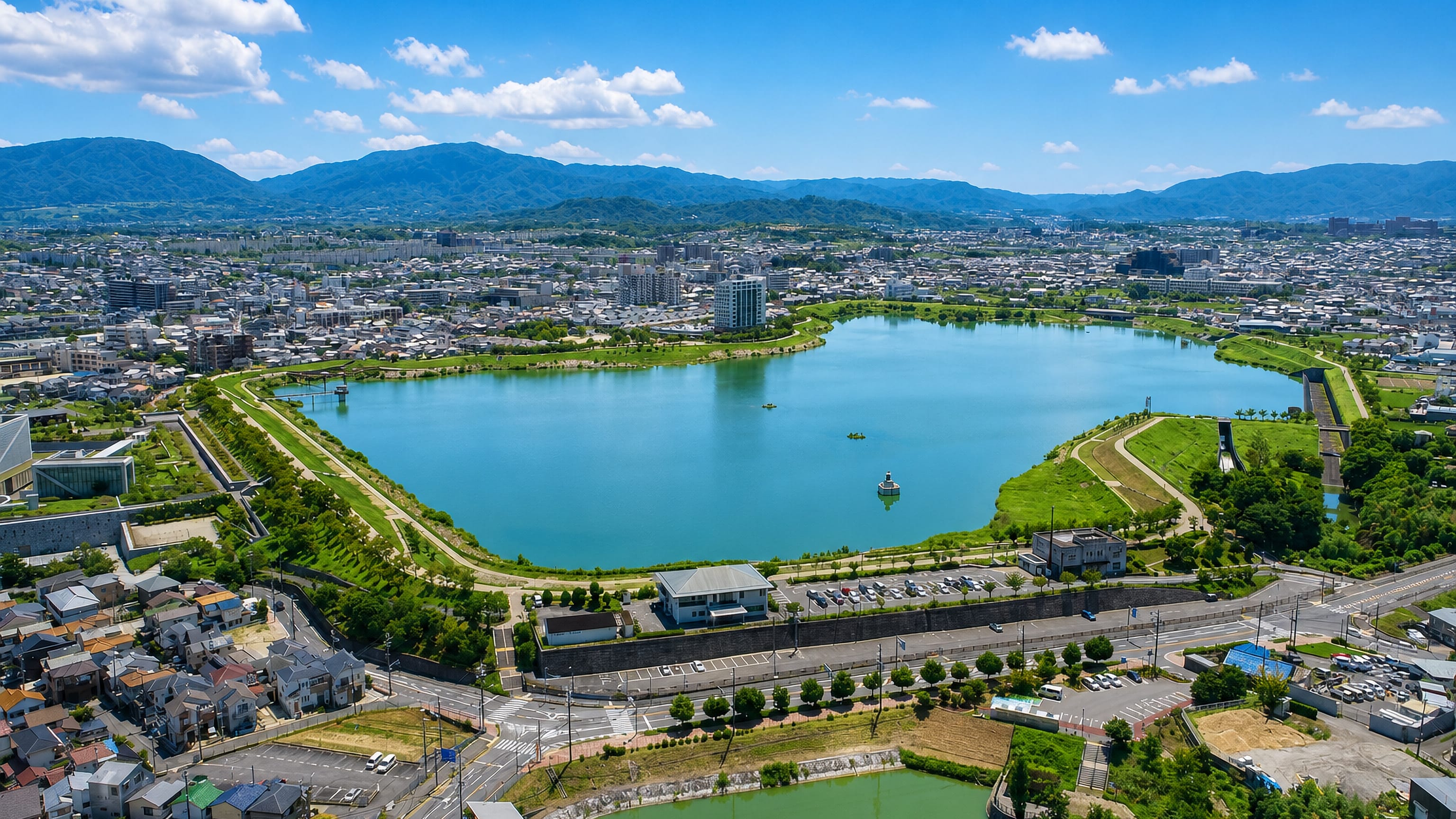 Aerial view of the Sayama reservoir ringed by the streets and rooftops of Sayama, Osaka, with mountains beyond.