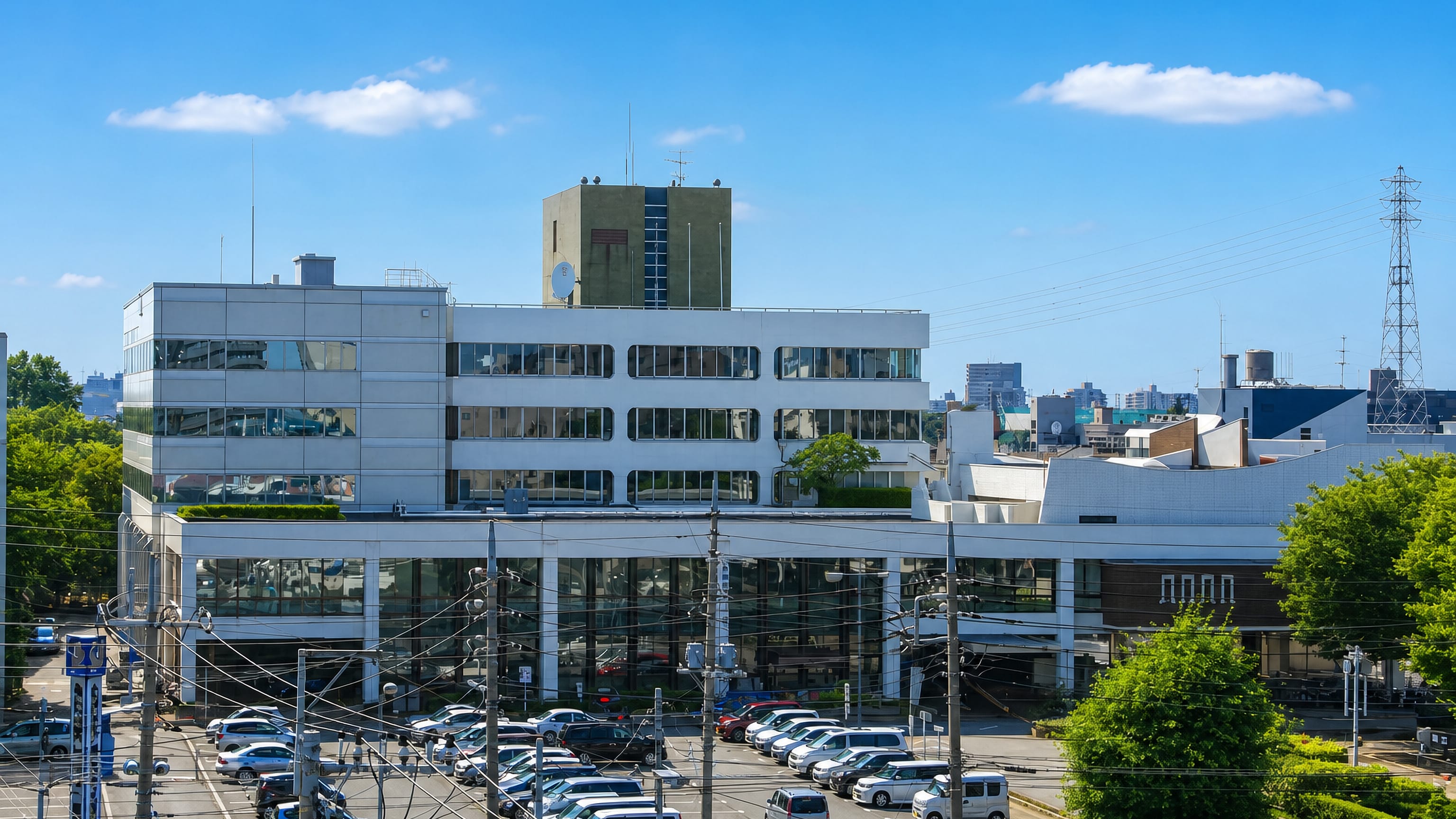 Mid-rise civic and office buildings above a busy car park under summer skies in Asaka, Saitama.