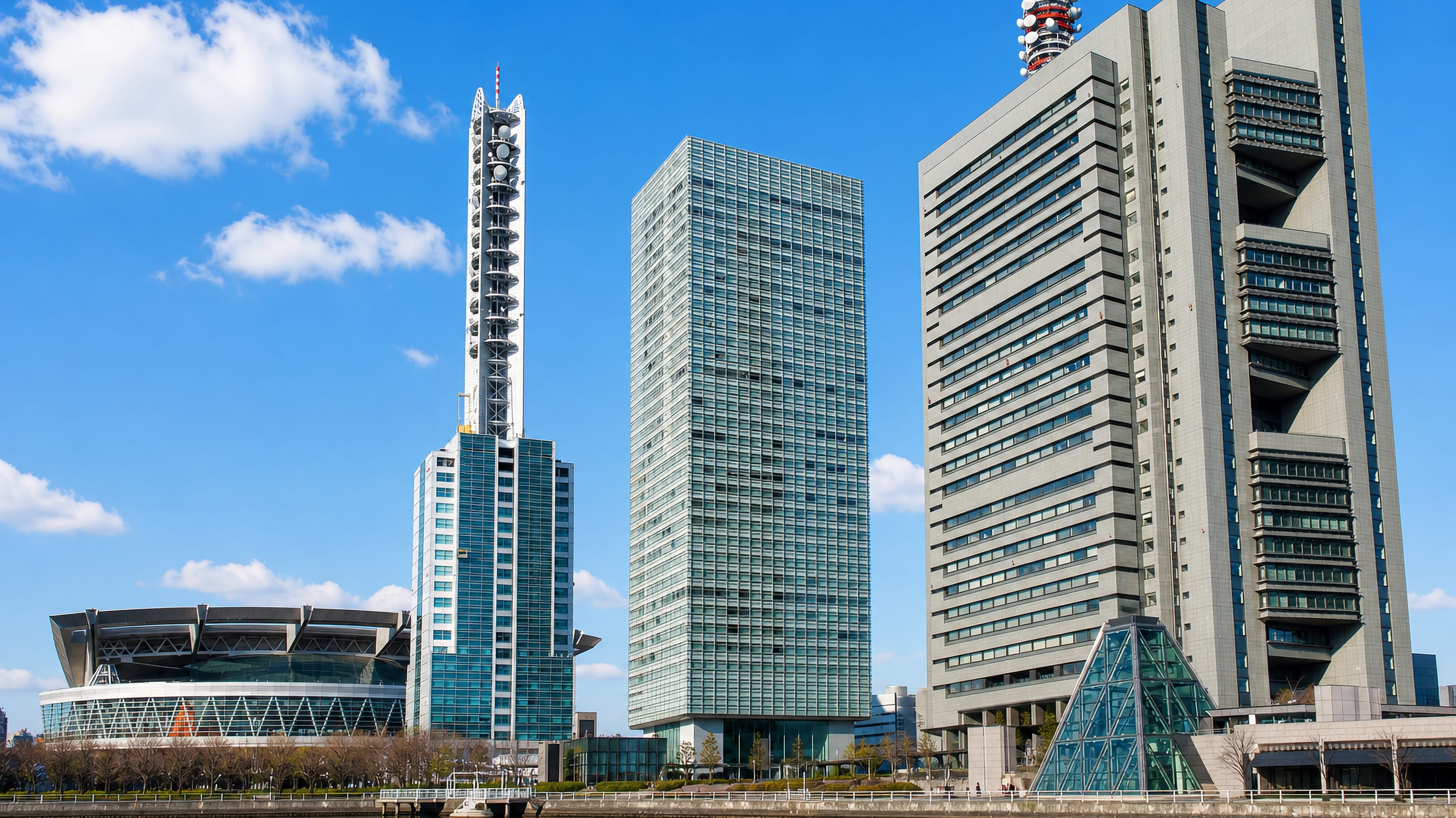 Glass towers, a broadcast mast and a domed arena rising above a waterfront promenade in Saitama, Japan.