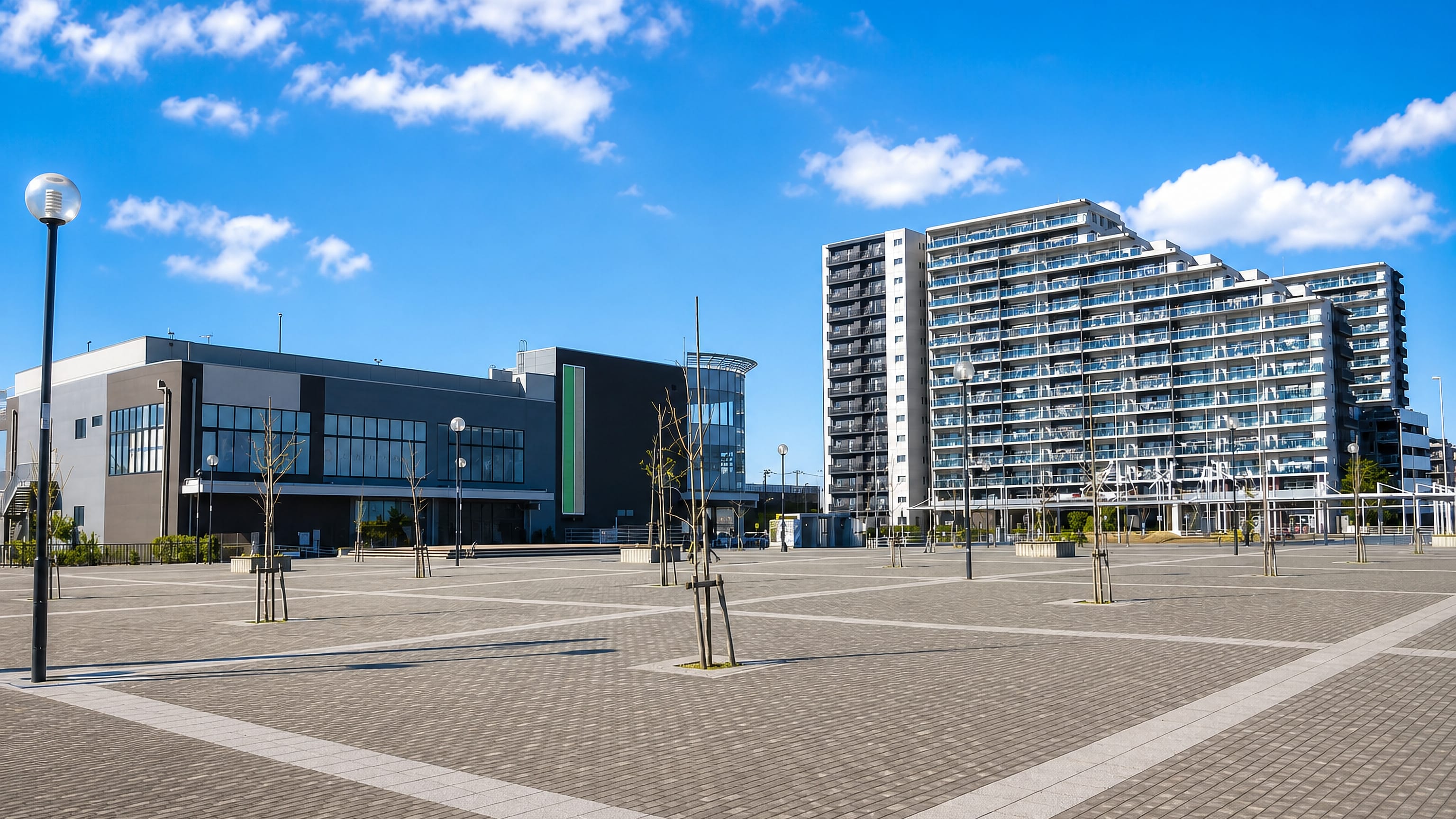 Wide paved station plaza in Yoshikawa, Saitama, with a modern hall and high-rise apartment blocks.