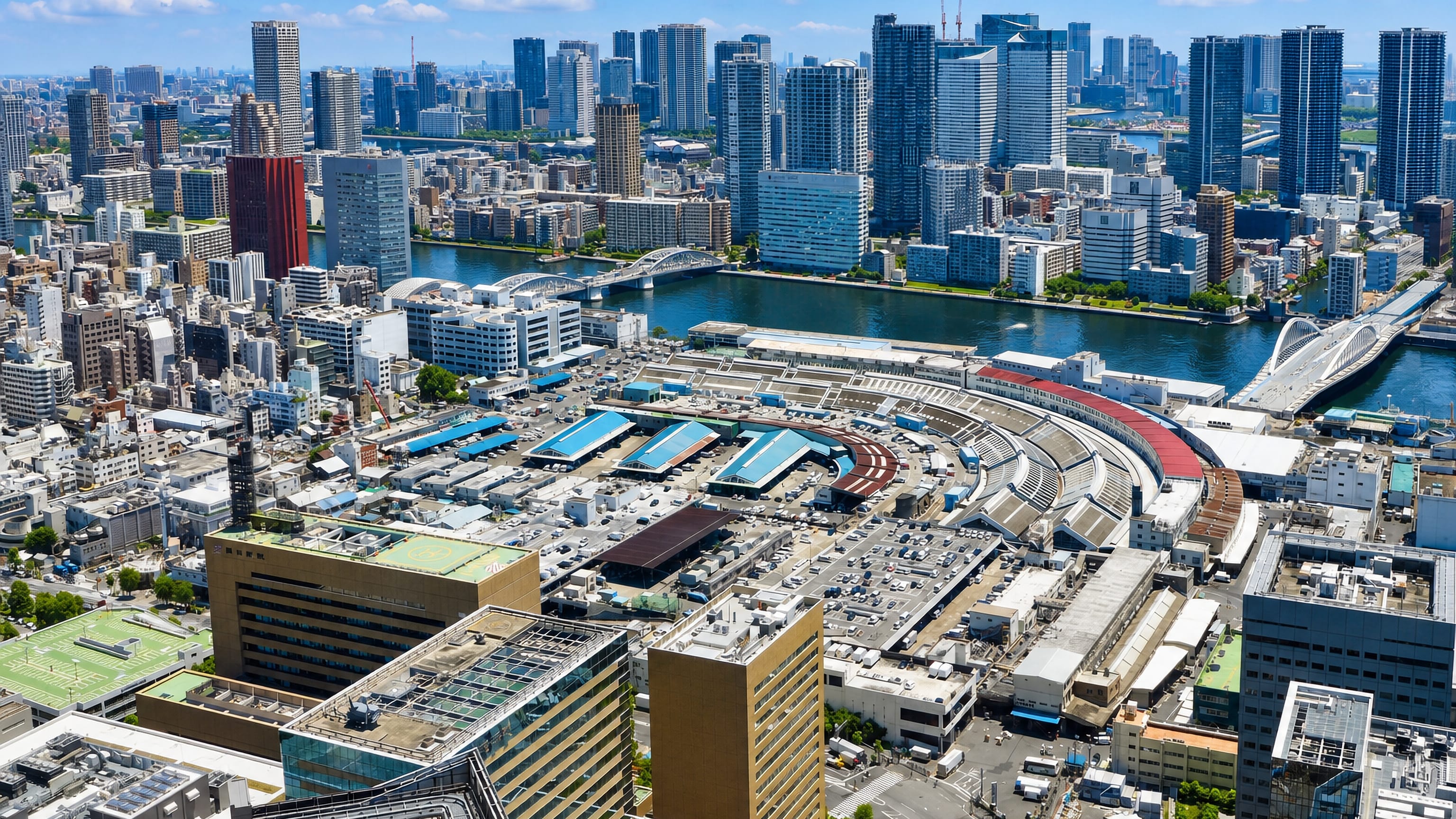Aerial of Tsukiji's curved market halls beside the Sumida River and Tokyo's Chuo skyline.