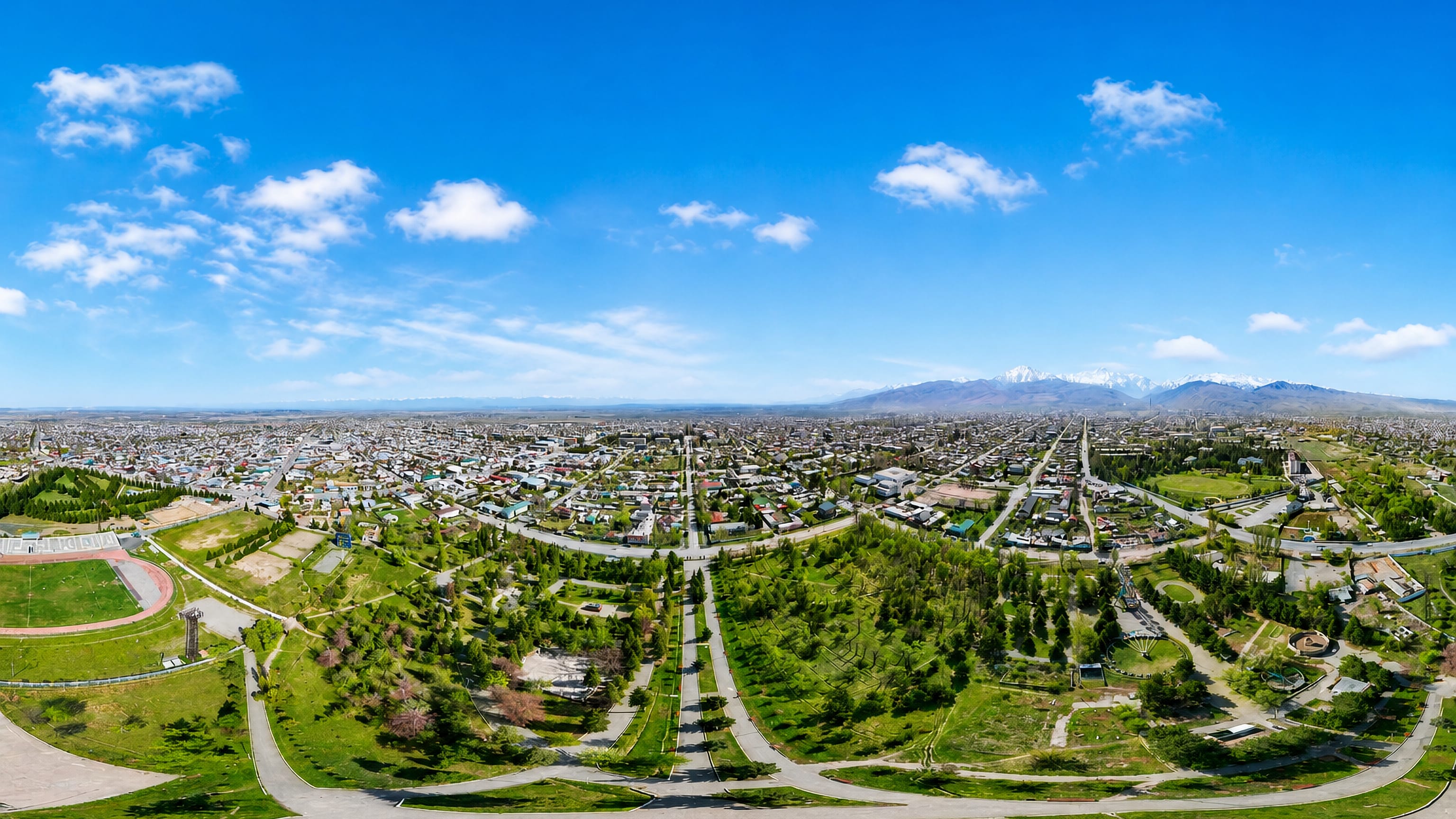 Aerial panorama of Karakol, Kyrgyzstan, its low-rise grid and parkland backed by snow-capped Tian Shan mountains.