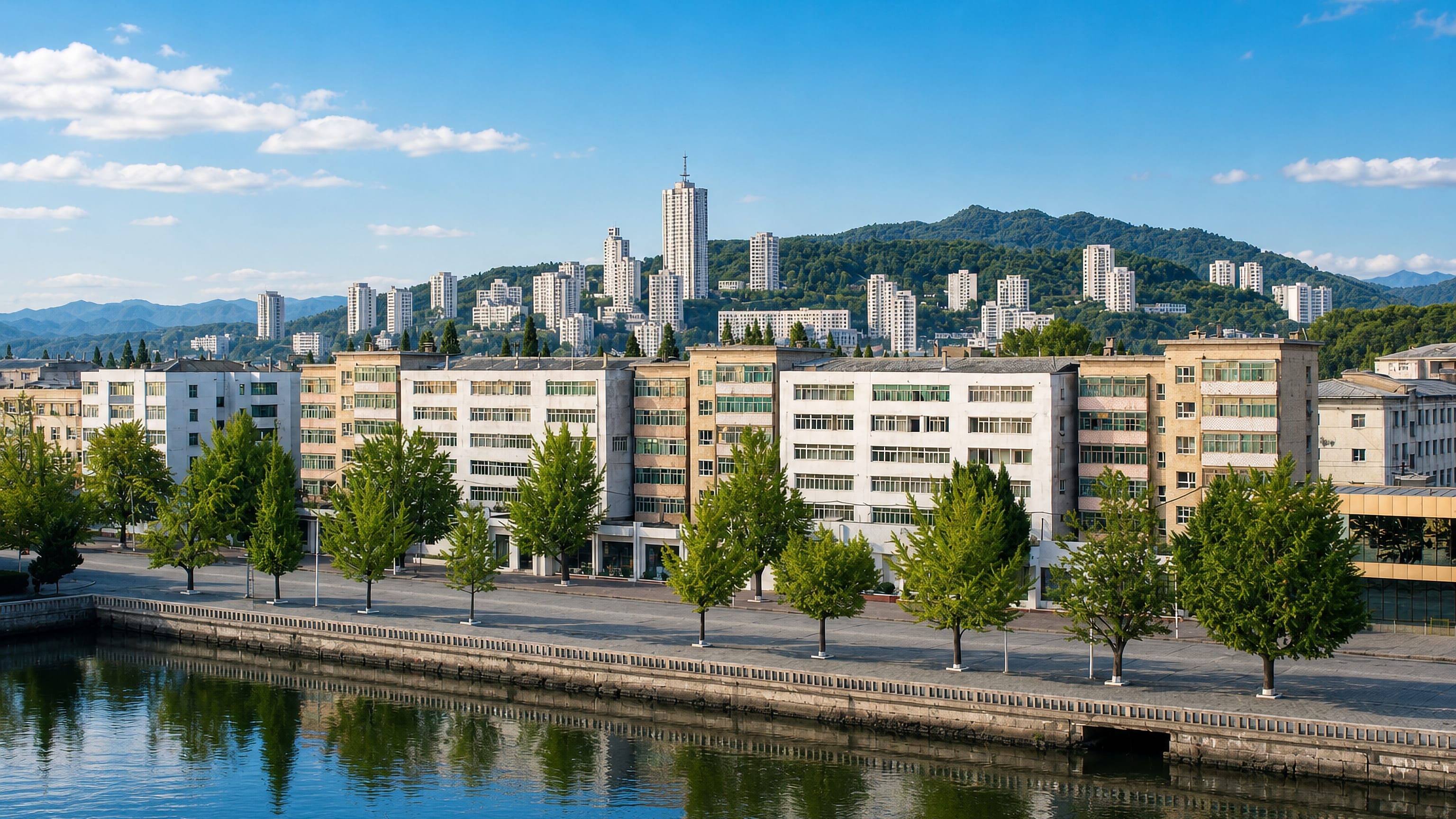 Tree-lined waterfront promenade and pastel apartment blocks below wooded hills in Wŏnsan, North Korea.
