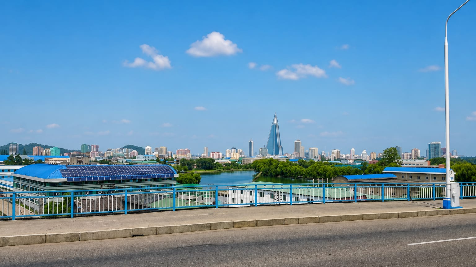 Pyongyang skyline across the Taedong River with the pyramid-shaped Ryugyong Hotel rising above the city under a blue sky.