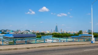 Pyongyang skyline across the Taedong River with the pyramid-shaped Ryugyong Hotel rising above the city under a blue sky.