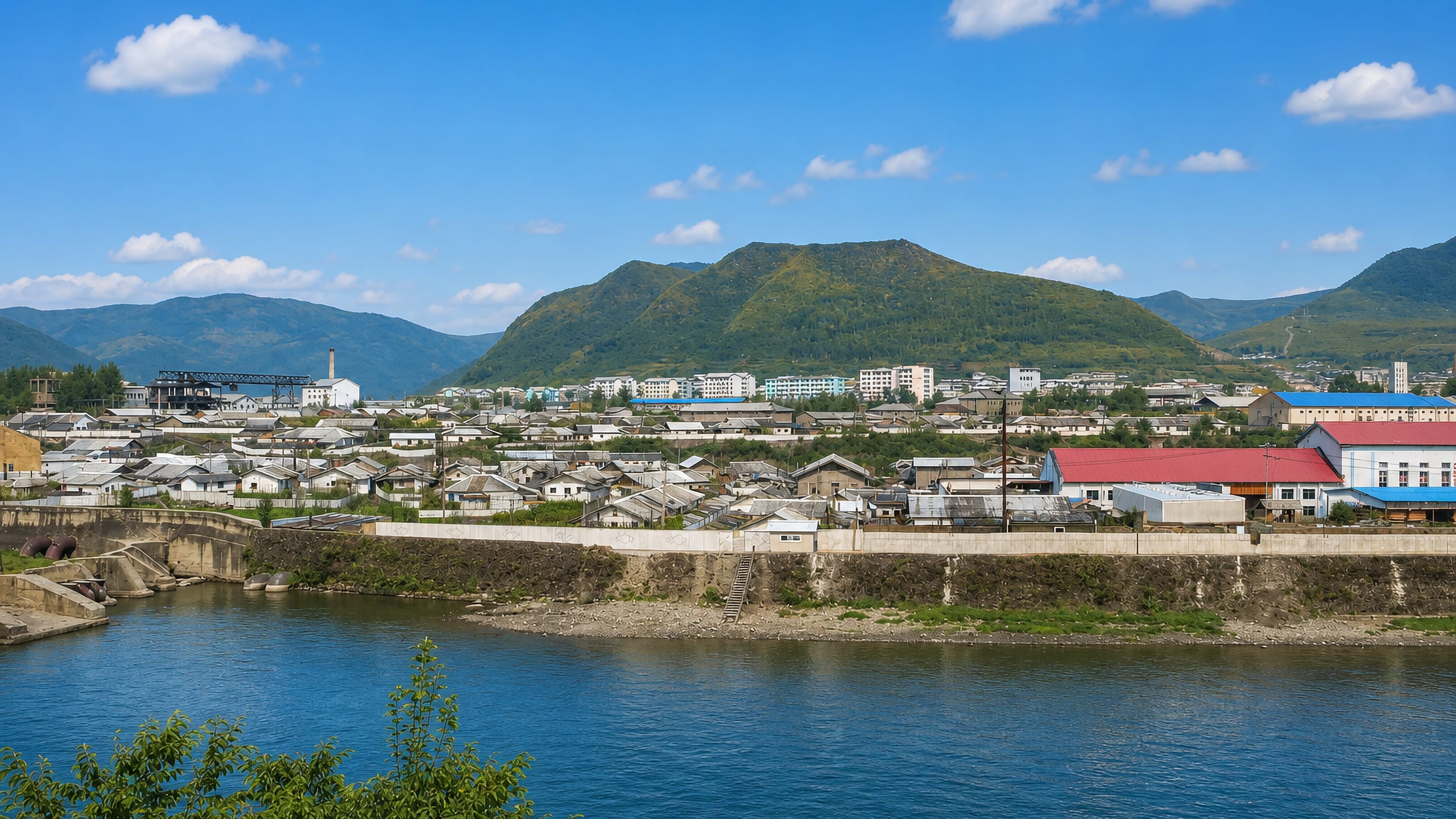 Hyesan, North Korea, low buildings and factory chimneys along the Yalu River beneath forested mountains.