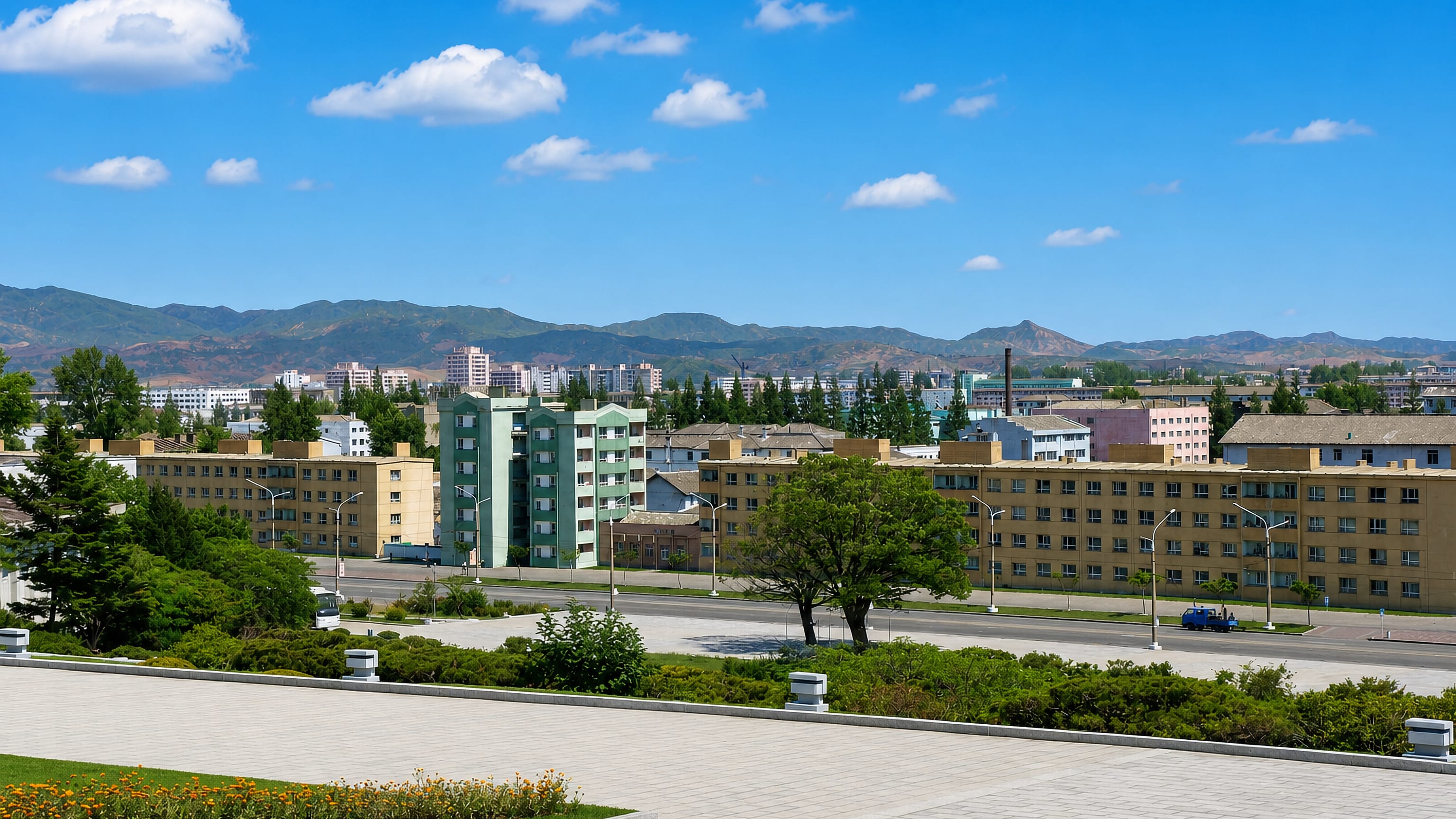 Low-rise apartment blocks and a wide boulevard in Hamhung, North Korea, backed by hazy green mountain ridges.