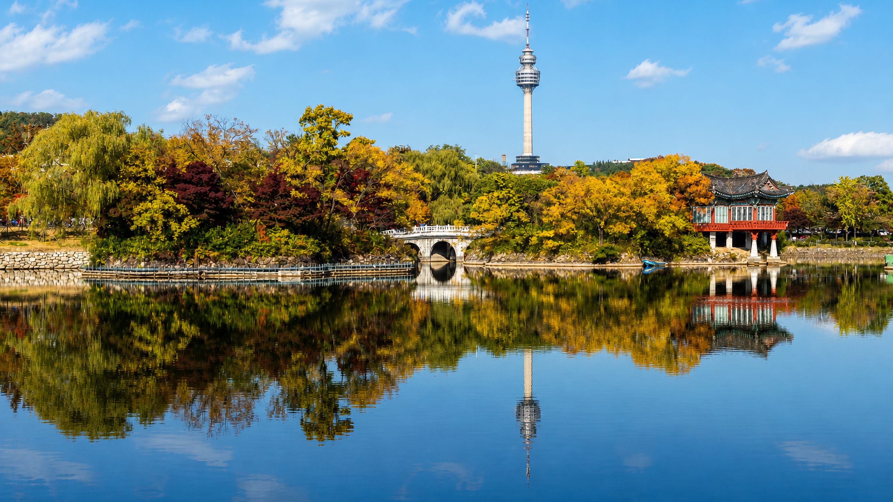 Daegu's 83 Tower above a lakeside park, with a traditional Korean pavilion and autumn foliage on the water.