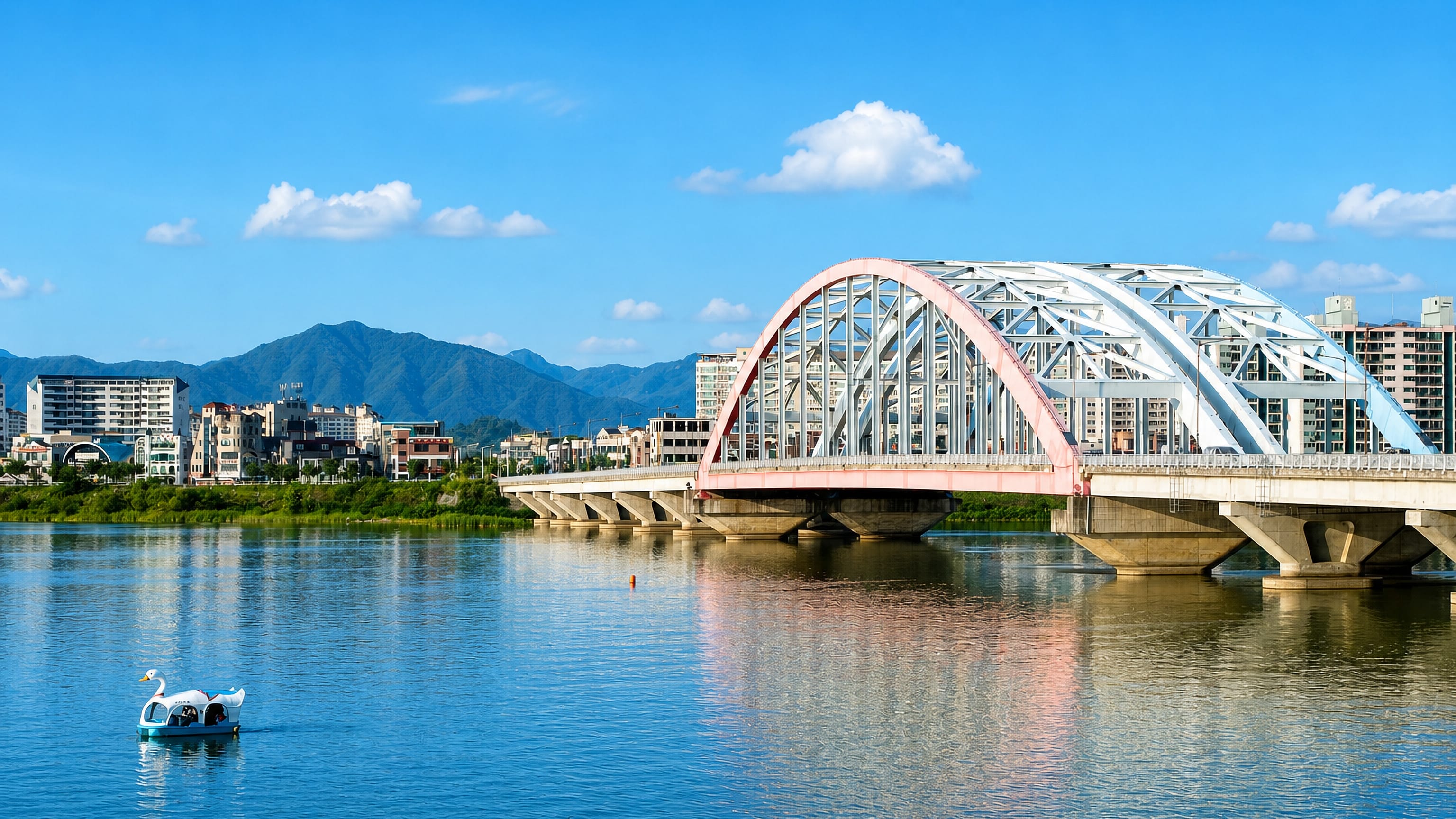 A pink steel arch bridge spanning the river at Chuncheon, with a swan pedal boat and green hills beyond