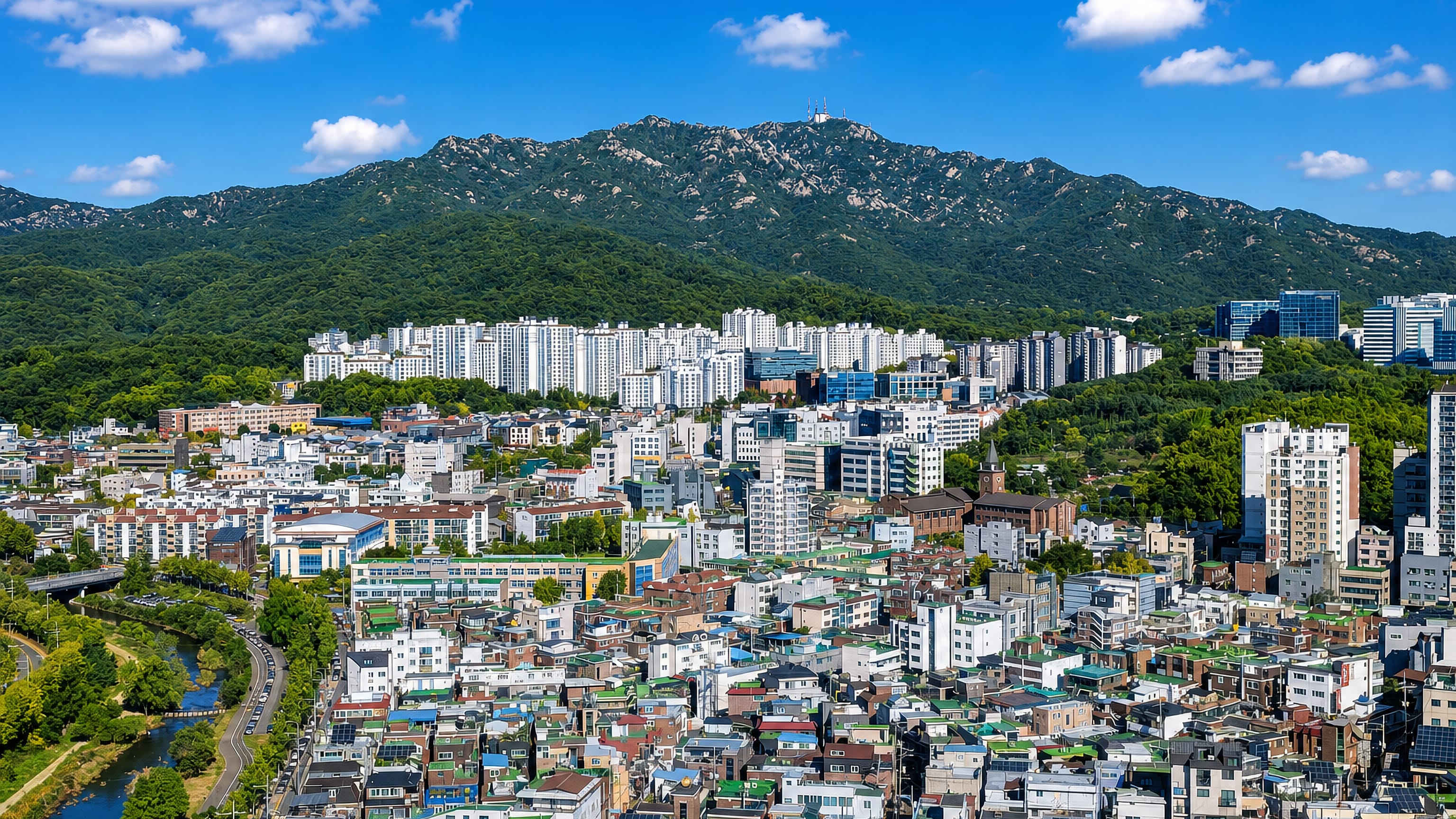 Apartment towers and low-rise streets of Anyang-si, South Korea, below a forested mountain ridge.