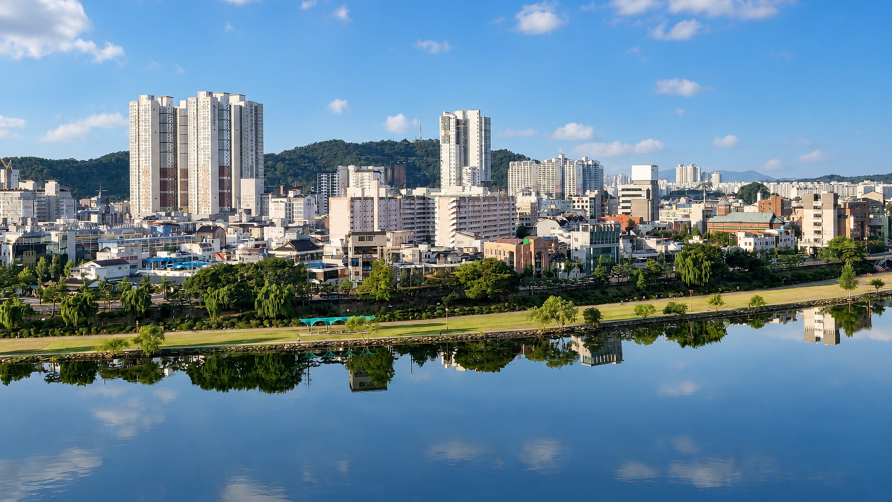 Chinju's riverside apartment towers and wooded hills mirrored in the calm Nam River under a bright summer sky.