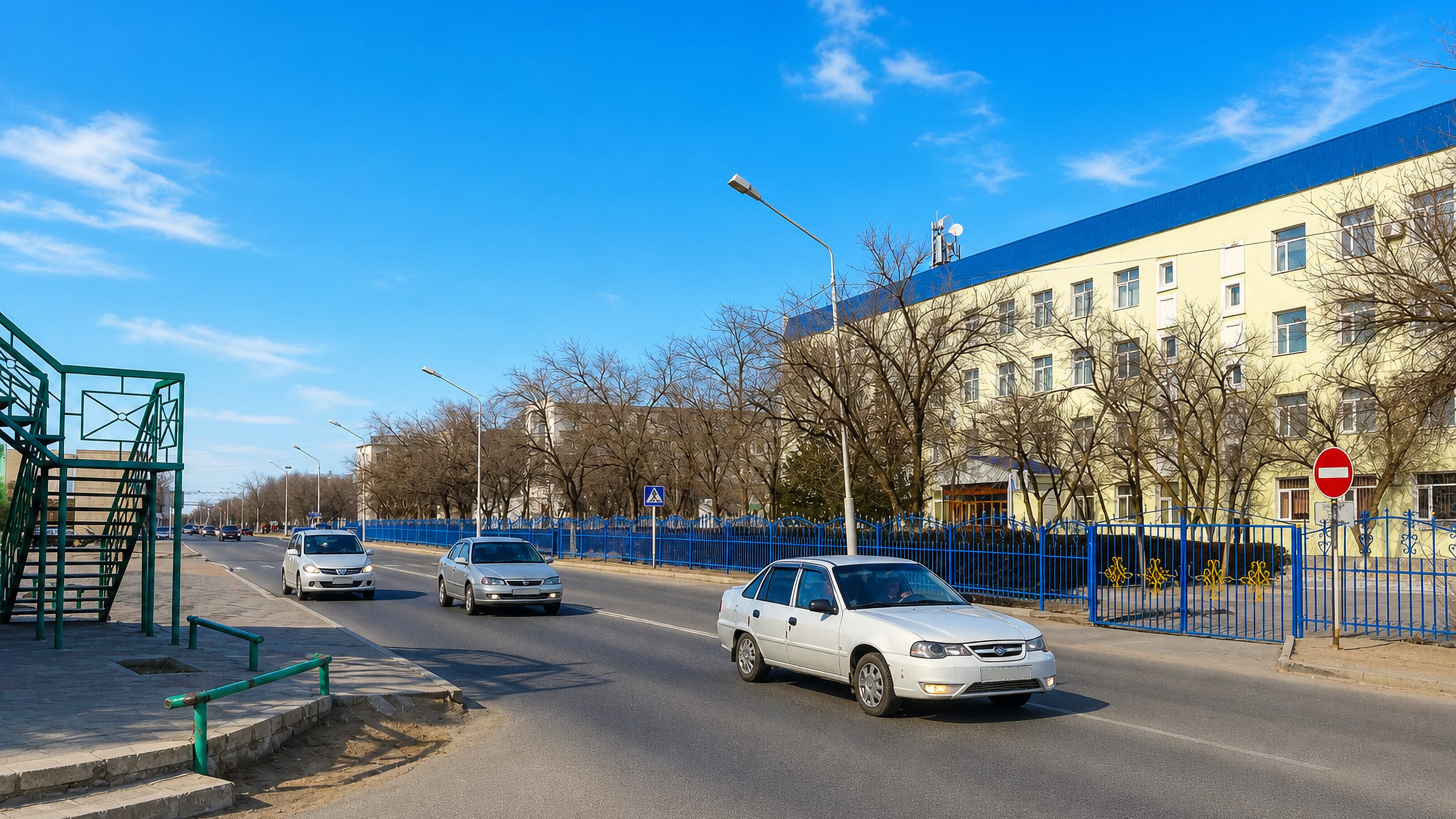 A wide sunlit avenue in Zhanaozen, Kazakhstan, lined with a pale four-storey block behind blue railings