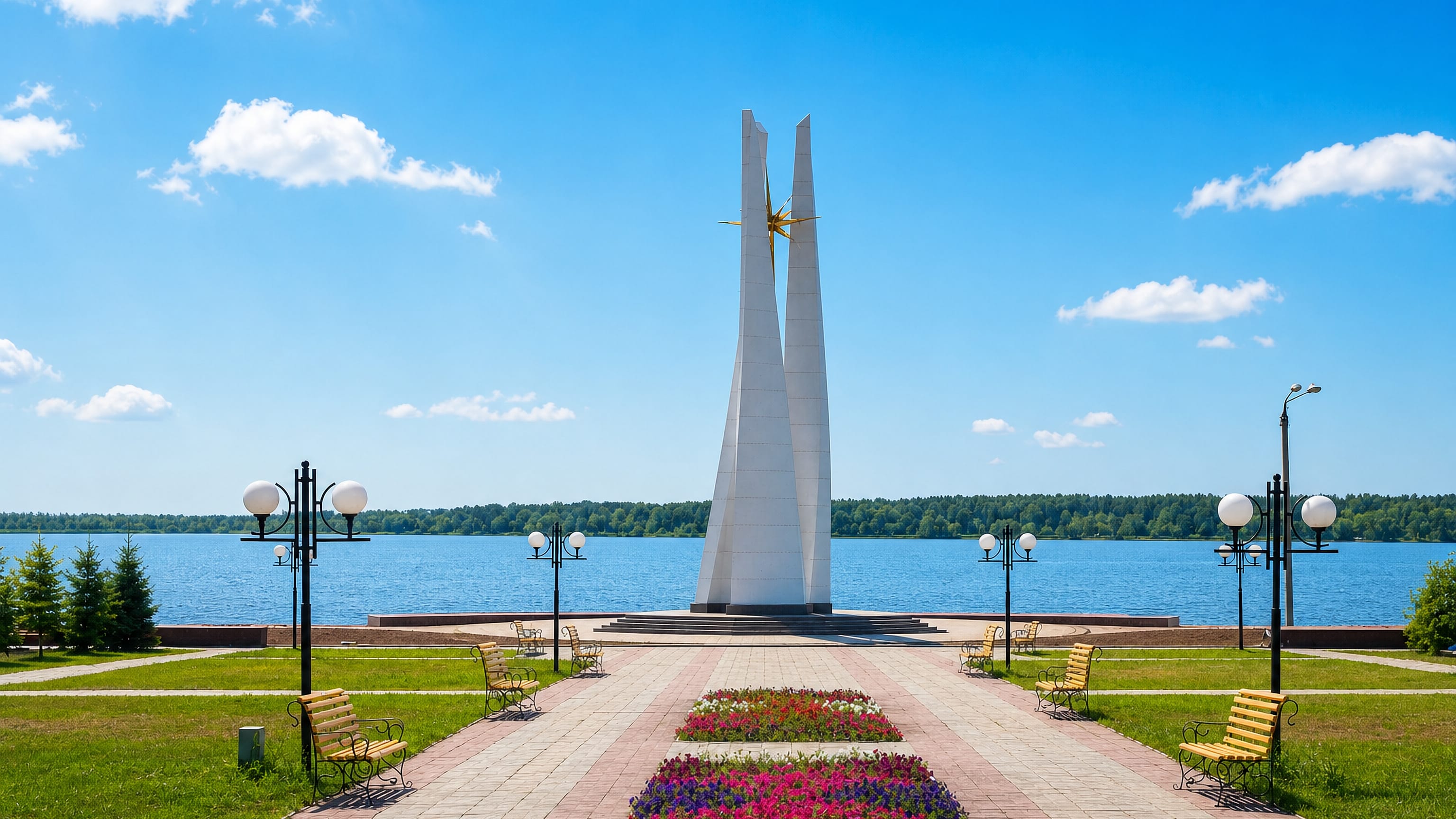 Waterside park in Petropavl, Kazakhstan, with a white twin-spire monument, flowerbeds and benches along the Ishim.