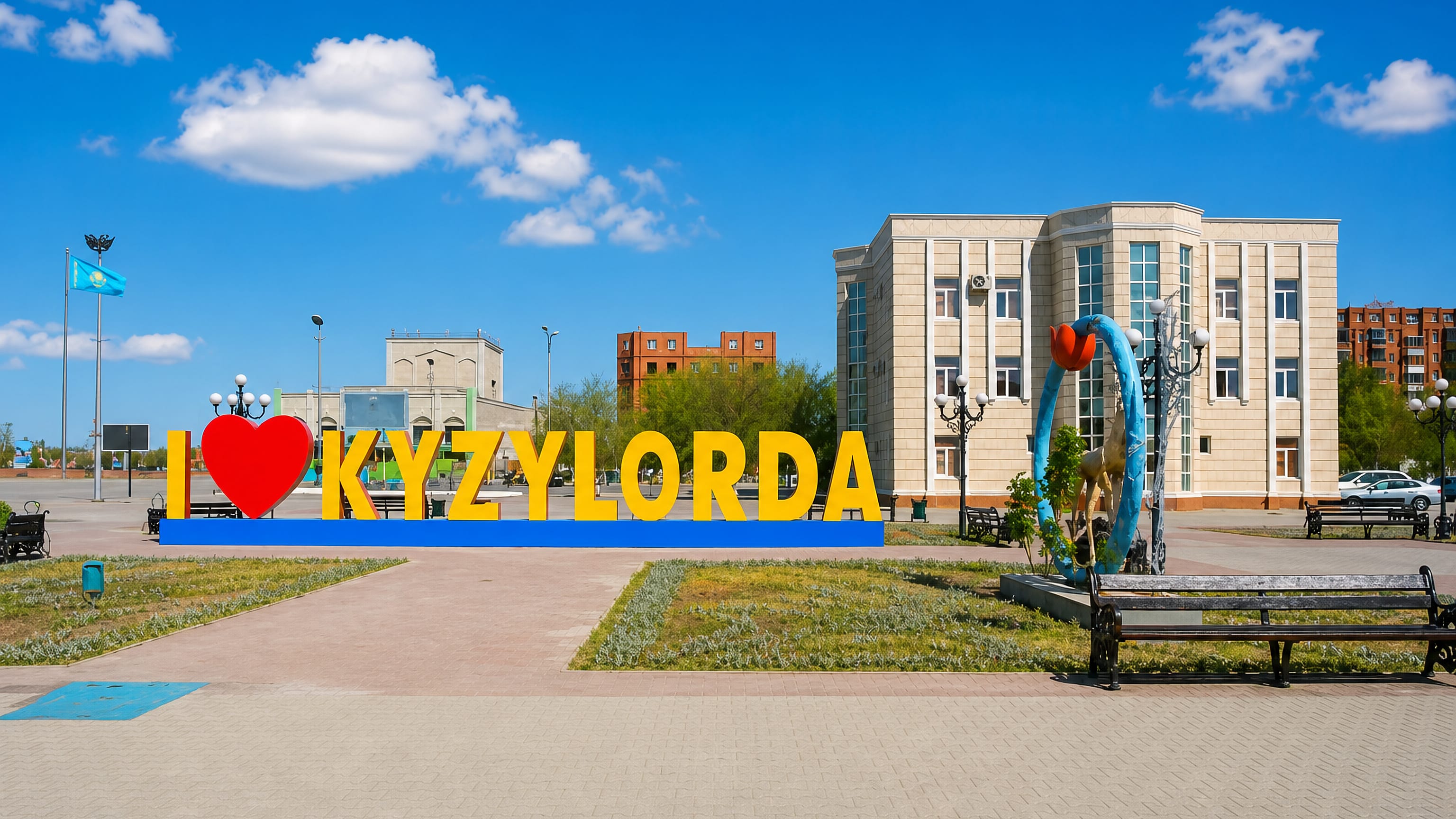 Central square in Kyzylorda, Kazakhstan, with the 'I love Kyzylorda' sign, flagpoles and a civic building.