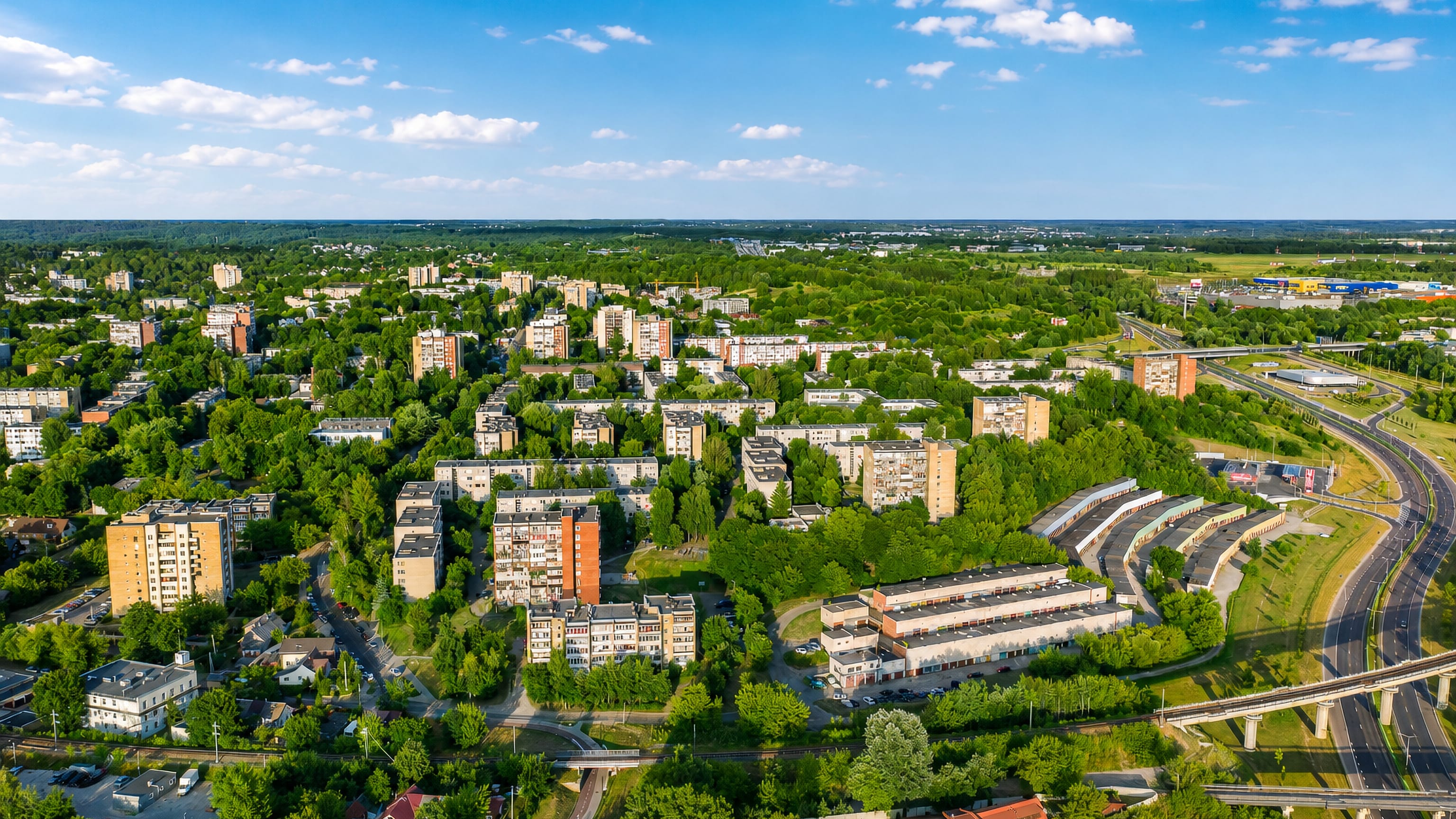 Aerial of Naujininkai's Soviet-era apartment blocks among trees beside Vilnius rail lines and bypass