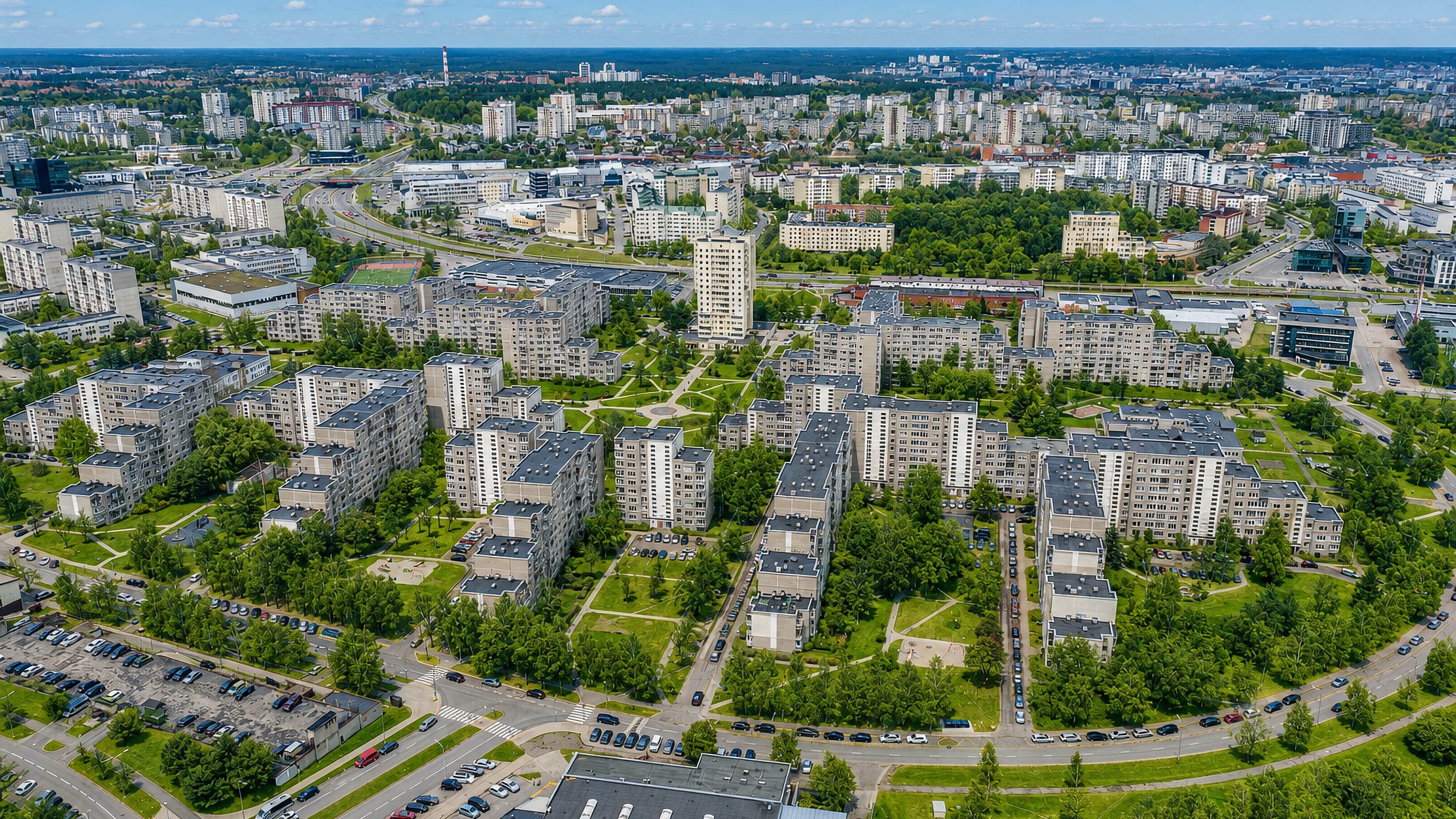Aerial view over Pašilaičiai in Vilnius, with mid-rise apartment blocks, tree-lined courtyards and distant city skyline.
