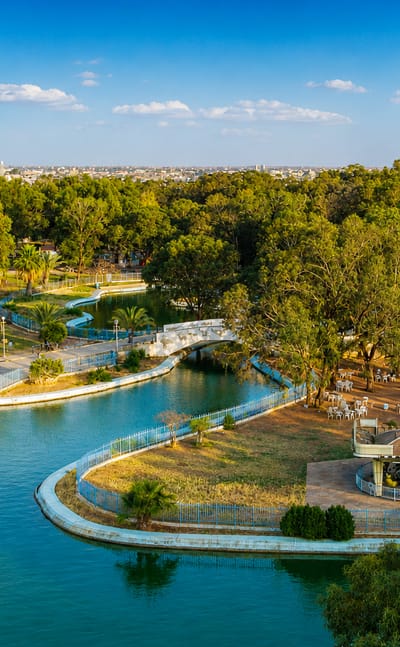 A lakeside city park with a bridge and gazebo backed by Benghazi's distant skyline, Libya