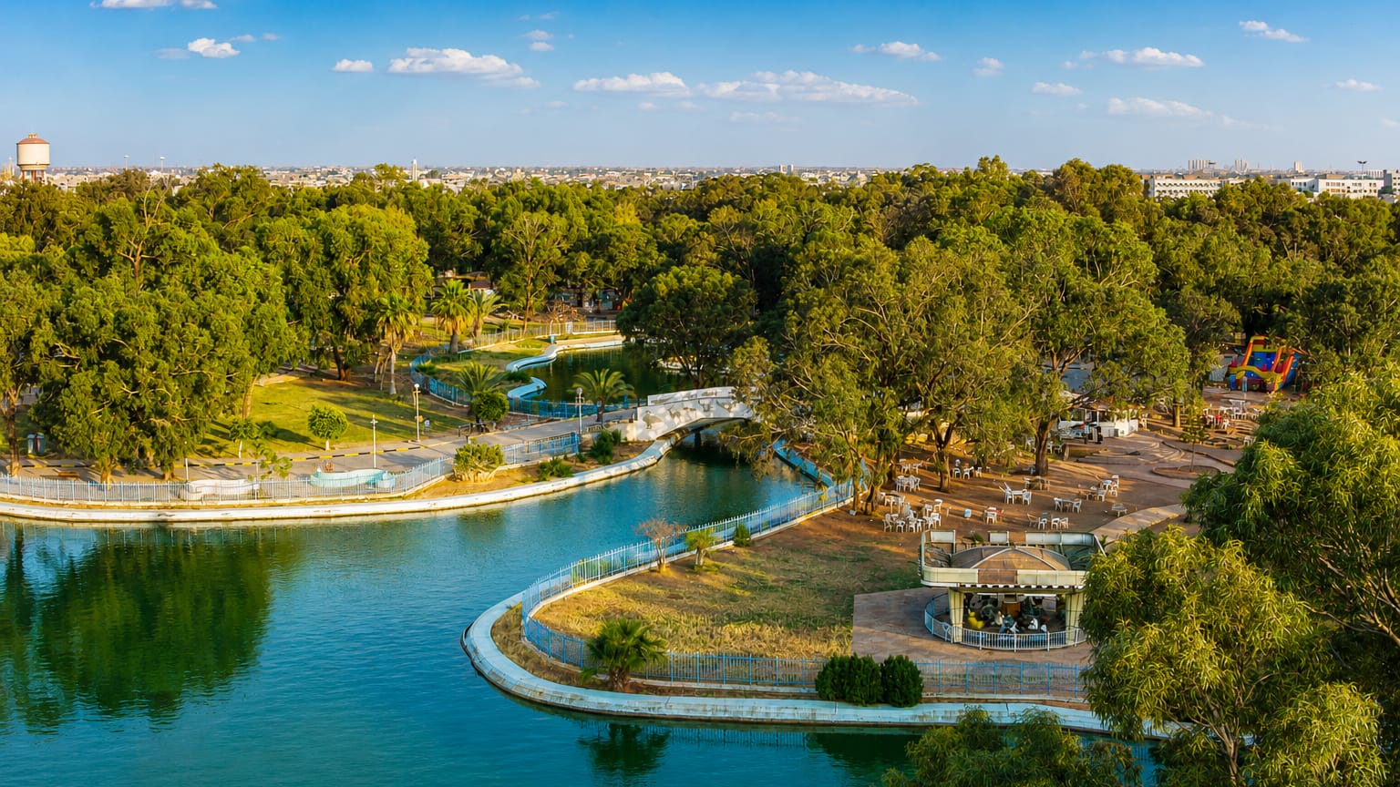A lakeside city park with a bridge and gazebo backed by Benghazi's distant skyline, Libya
