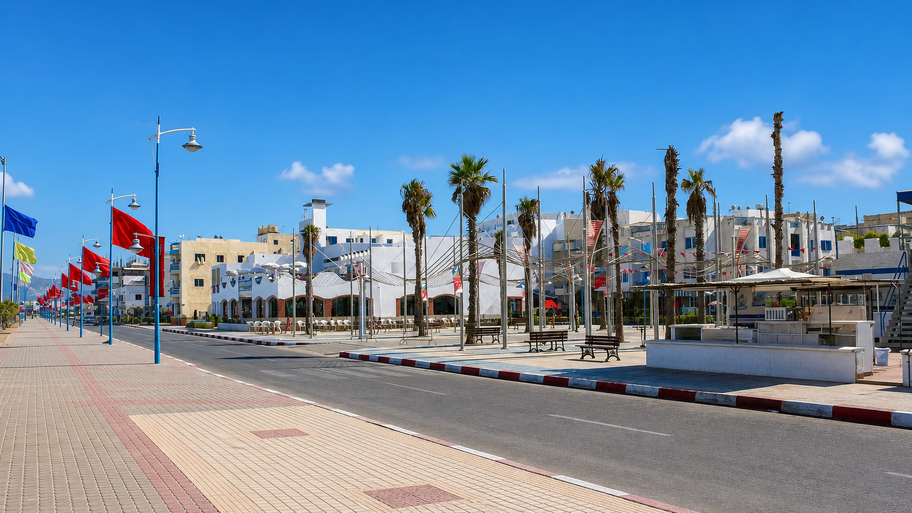Palm-lined seafront boulevard hung with Moroccan flags beside whitewashed buildings in Martil, Morocco.