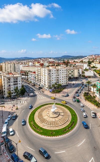 Aerial view of a Tetouan fountain roundabout, palm-lined plaza and white buildings with hills and sea beyond