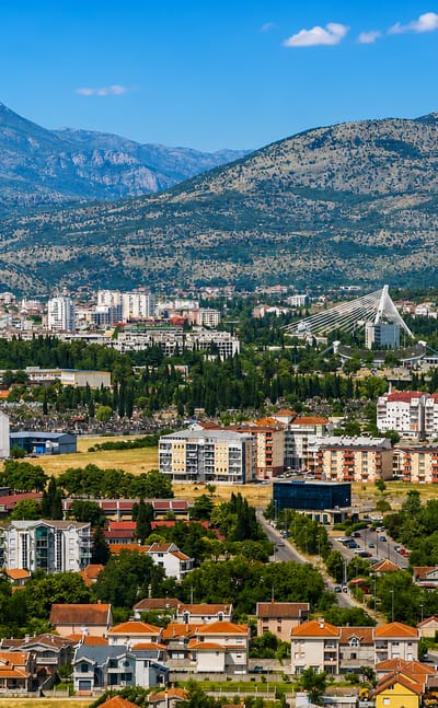 Podgorica in daylight, red-roofed houses and apartment blocks on the plain below rugged, partly snow-capped mountains.
