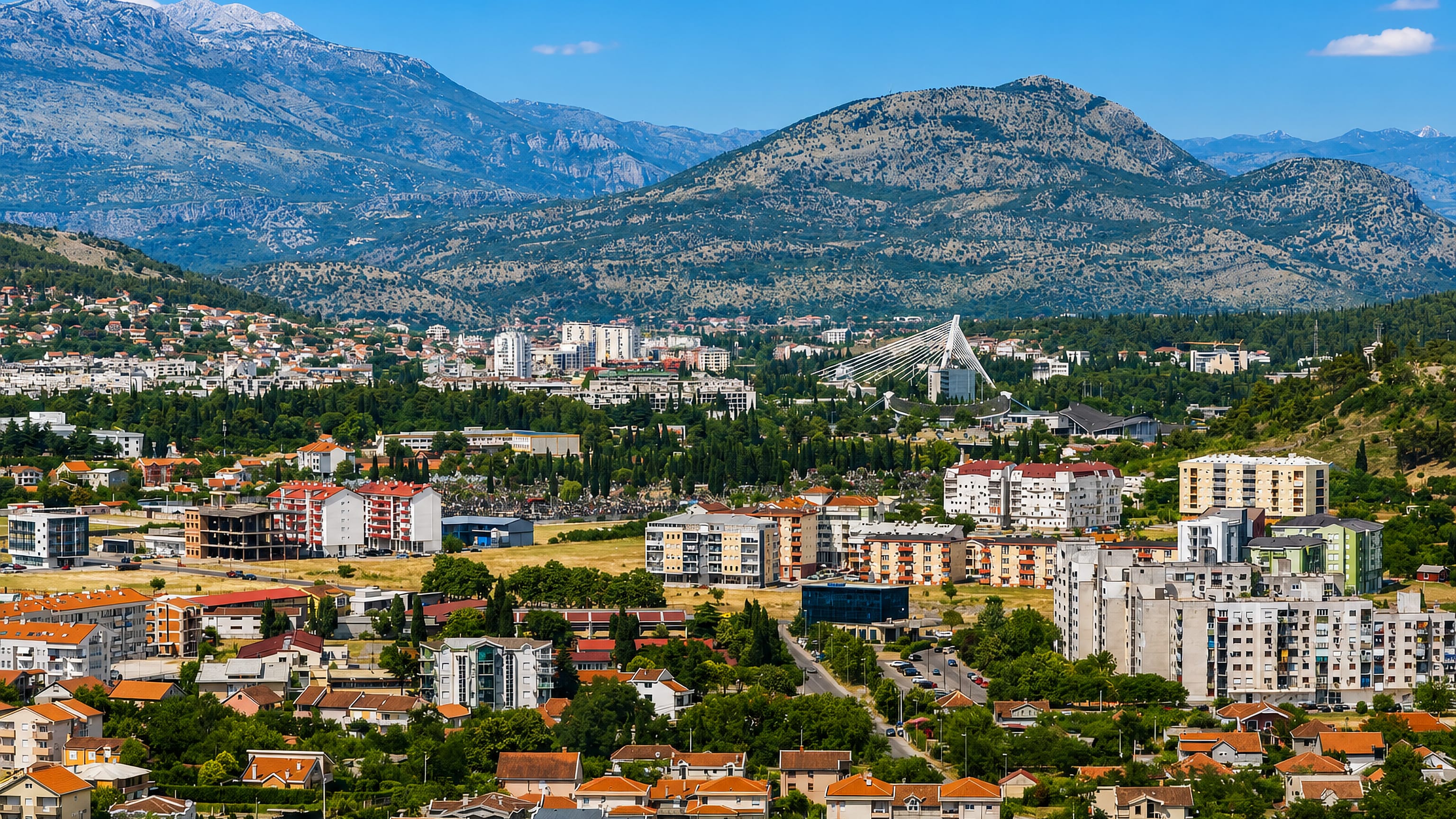 Podgorica in daylight, red-roofed houses and apartment blocks on the plain below rugged, partly snow-capped mountains.