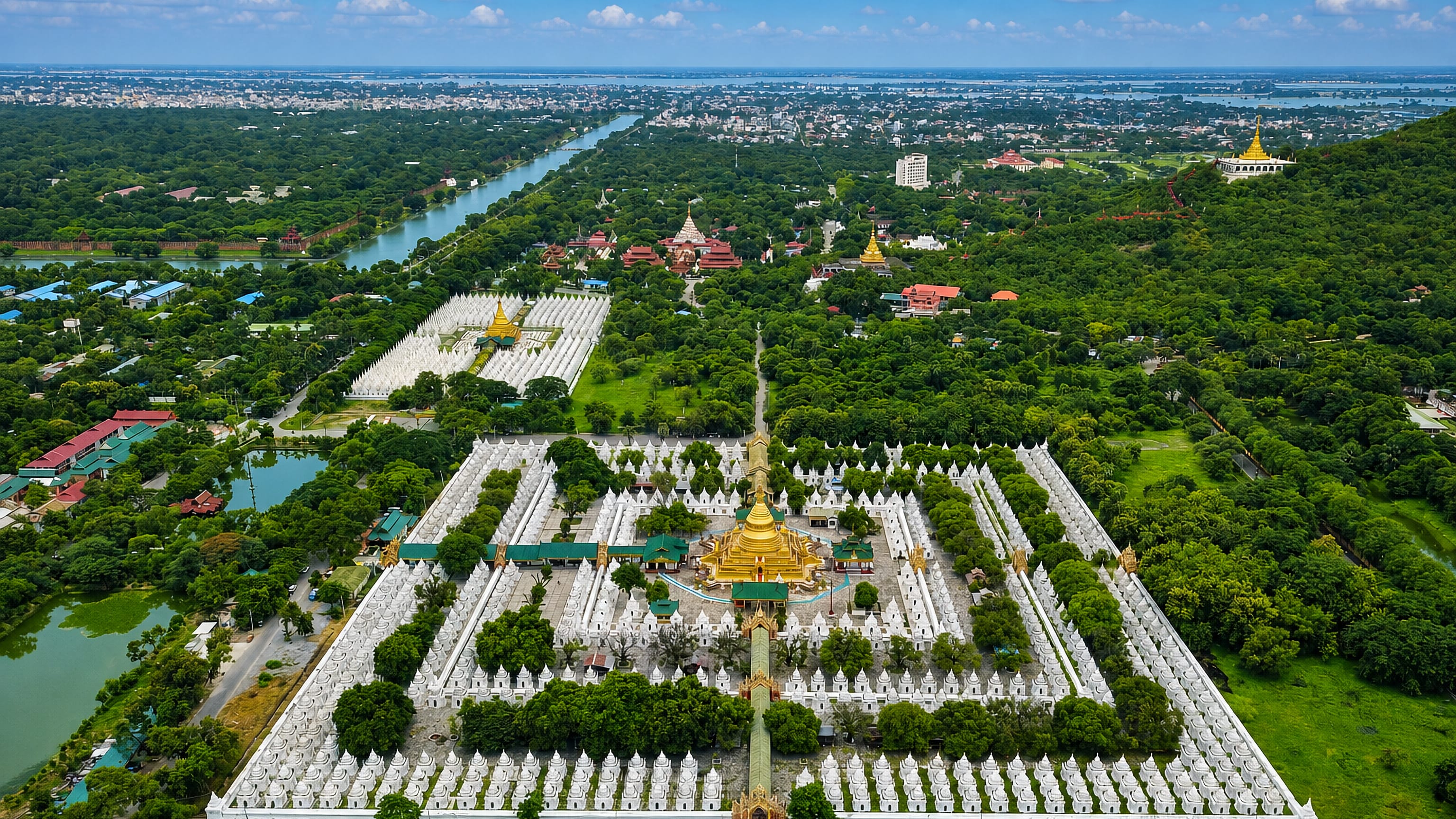 Aerial view of Mandalay's Kuthodaw Pagoda, rows of white stupas around a golden shrine, city beyond.