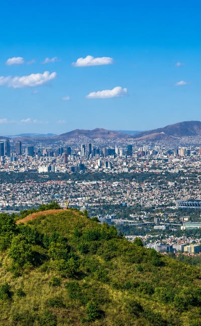 Panoramic view of Mexico City sprawling below the forested hills of Tlalpan, with distant mountains.
