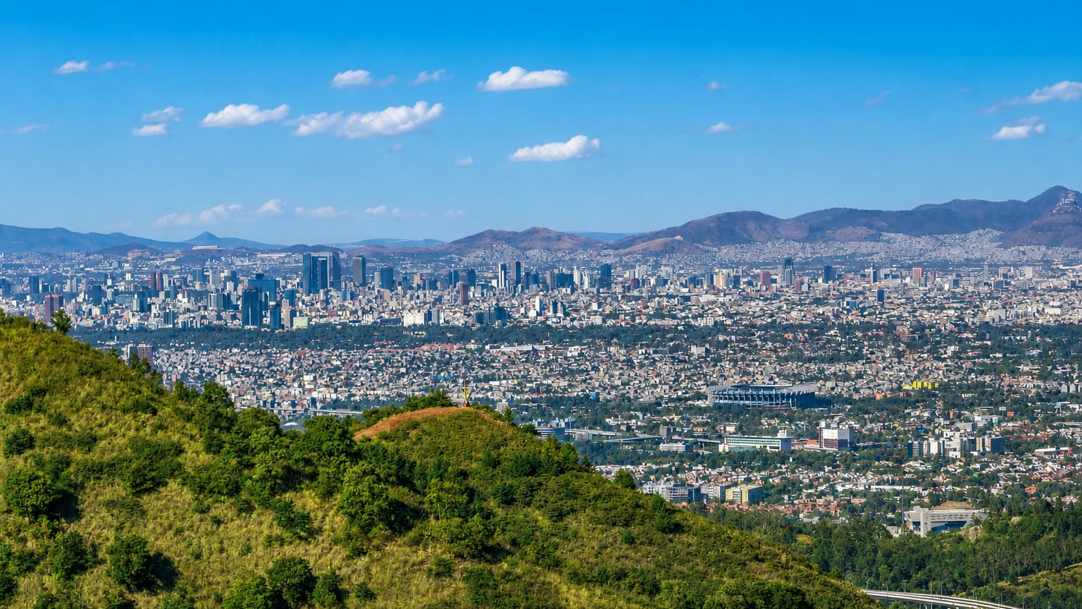 Panoramic view of Mexico City sprawling below the forested hills of Tlalpan, with distant mountains.