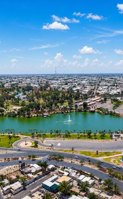Aerial view of Mexicali's flat sprawl around a park lake and a giant Mexican flag on a monumental flagpole.