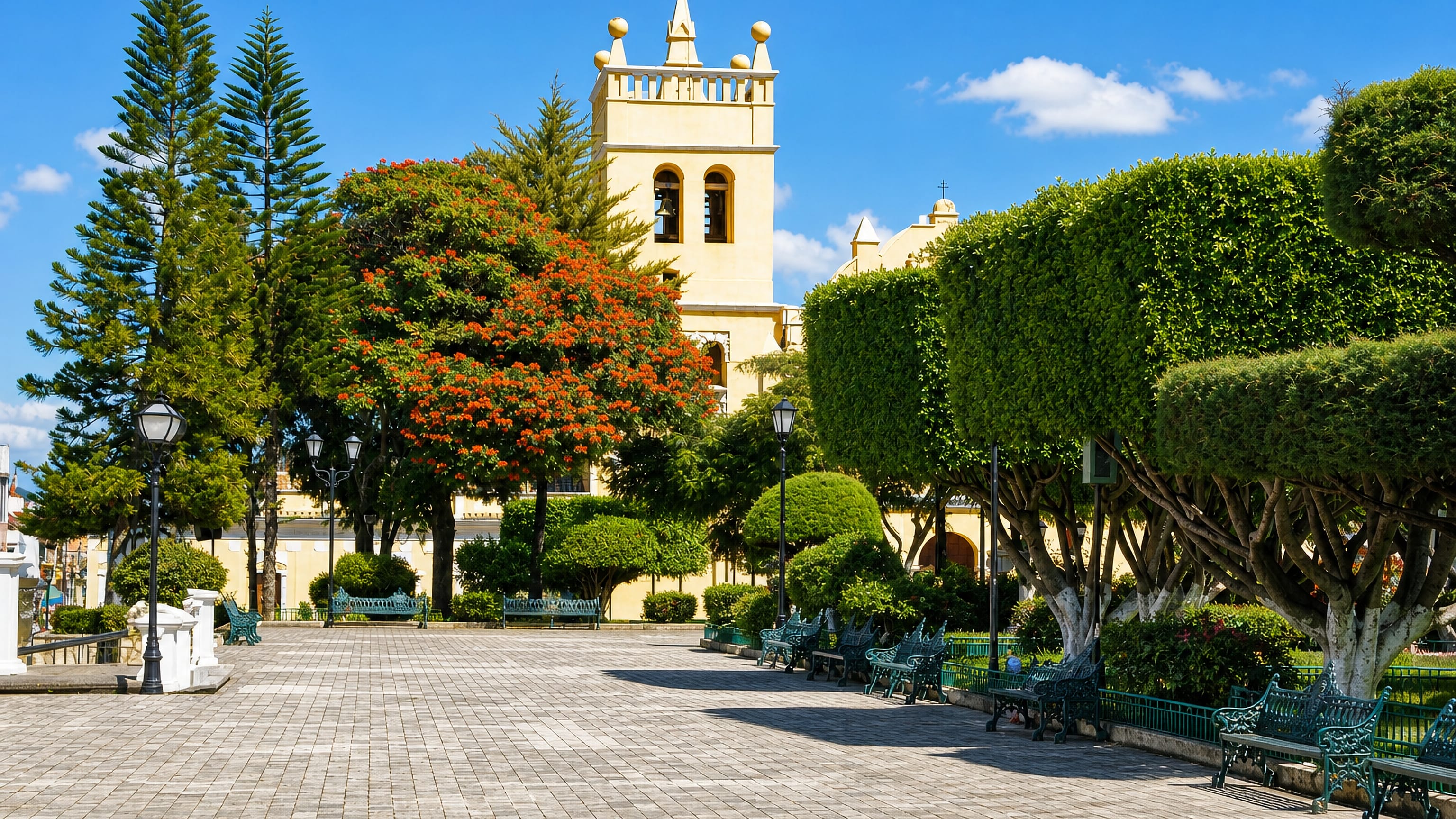 Paved central plaza with green benches, a flowering flame tree and a yellow church tower in Comitán, Chiapas.