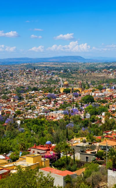 Hilltop view of San Miguel de Allende with the pink Parroquia spires above colourful rooftops and jacarandas.