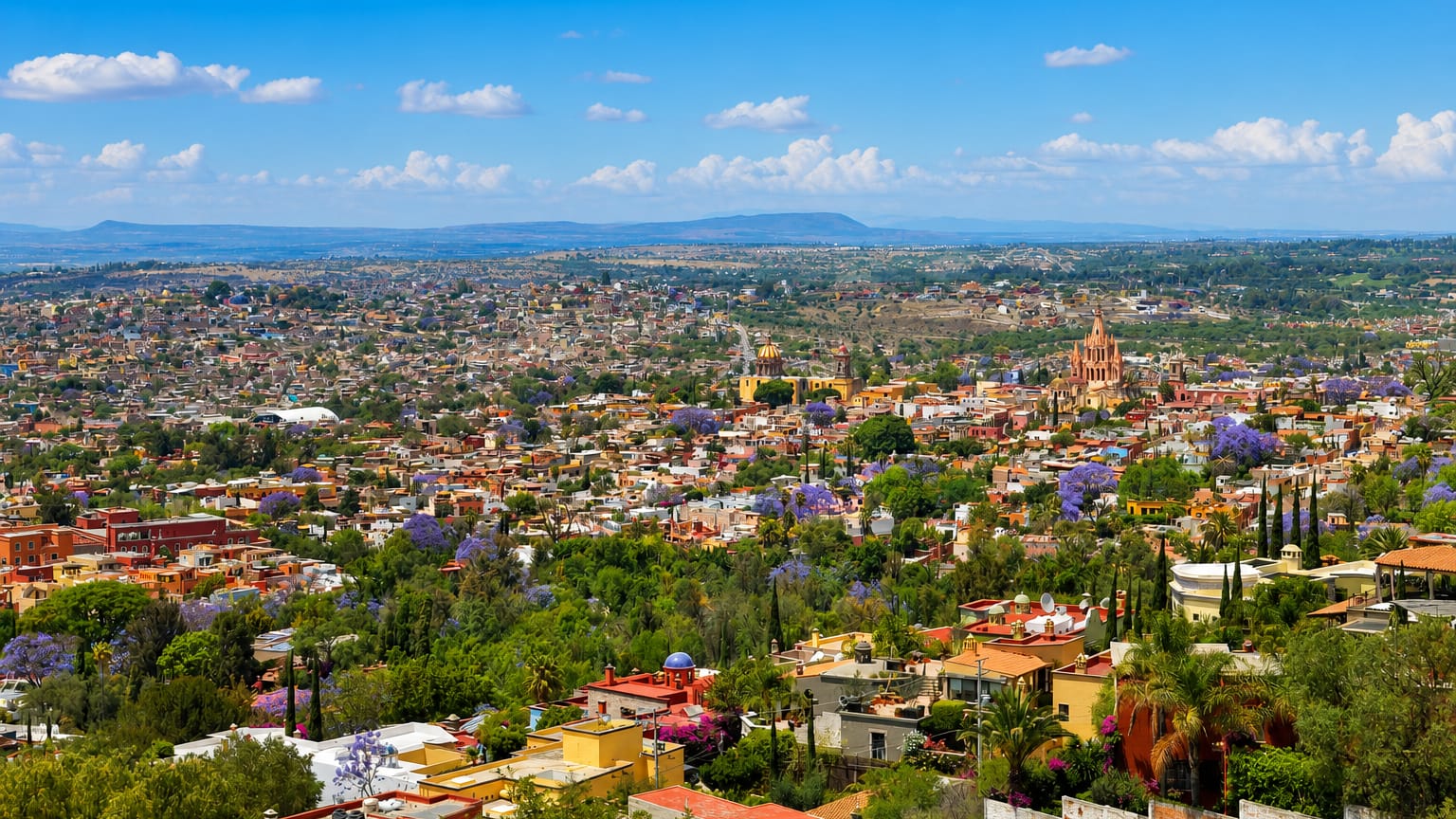 Hilltop view of San Miguel de Allende with the pink Parroquia spires above colourful rooftops and jacarandas.