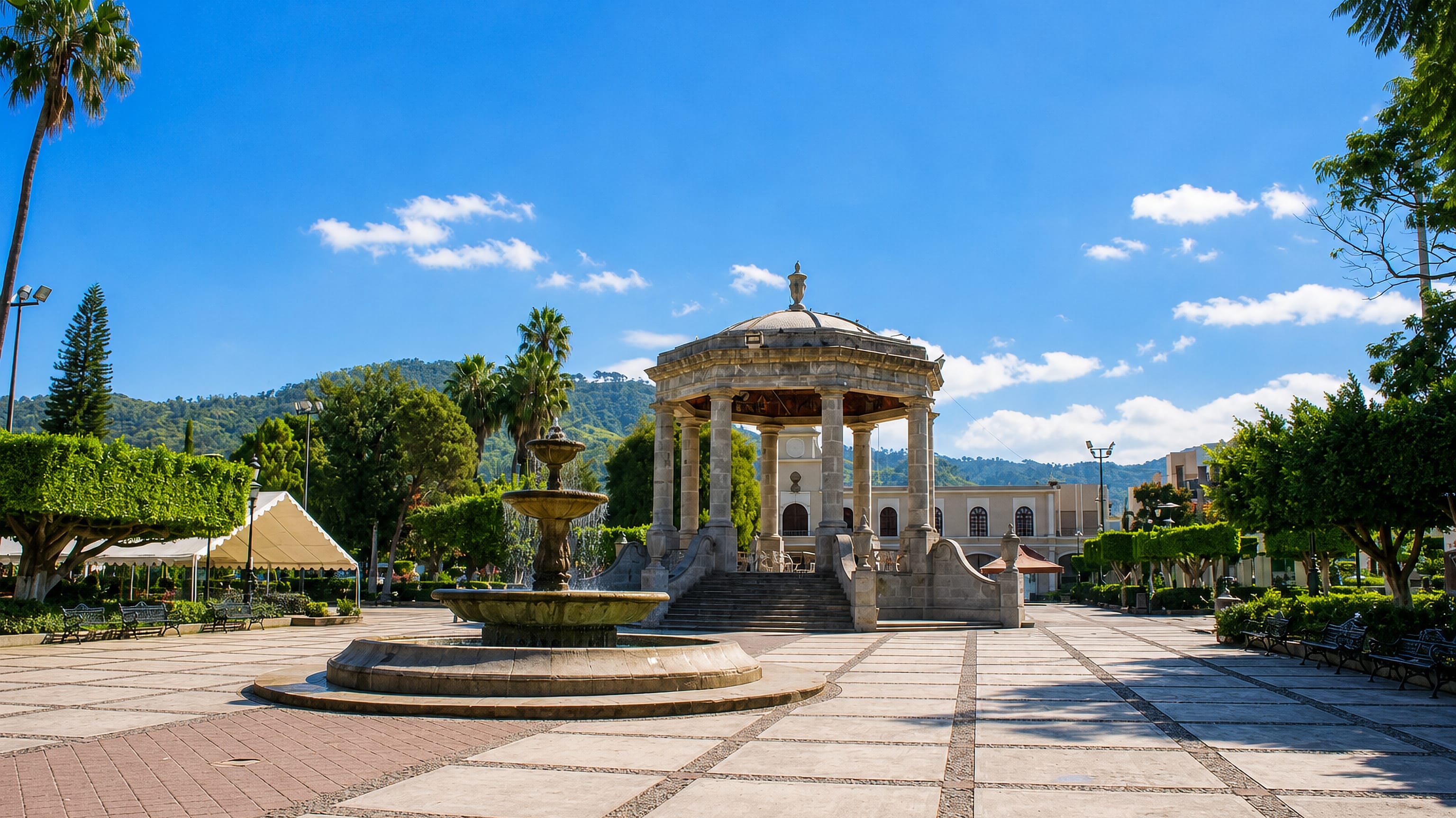 A stone rotunda bandstand and fountain on the tiled main plaza of Ciudad Guzmán, Jalisco, with green hills behind.