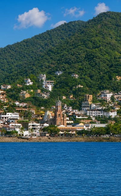 Waterfront view of Puerto Vallarta with the crowned Guadalupe church tower below jungle-covered hills.
