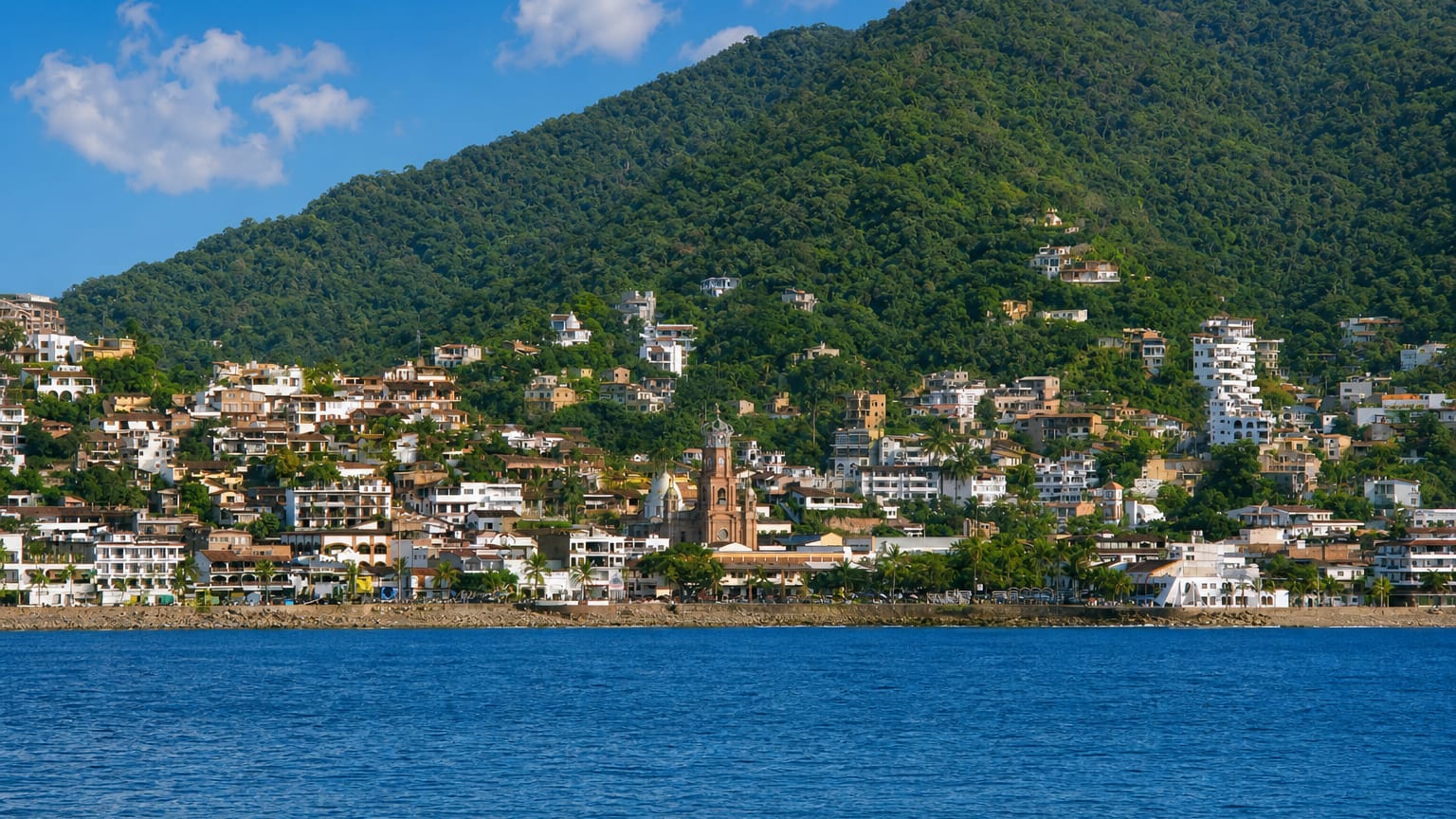 Waterfront view of Puerto Vallarta with the crowned Guadalupe church tower below jungle-covered hills.