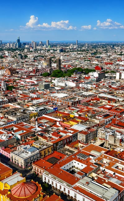 Aerial view of Puebla's colonial centre, red-tiled rooftops and cathedral towers with distant skyscrapers.