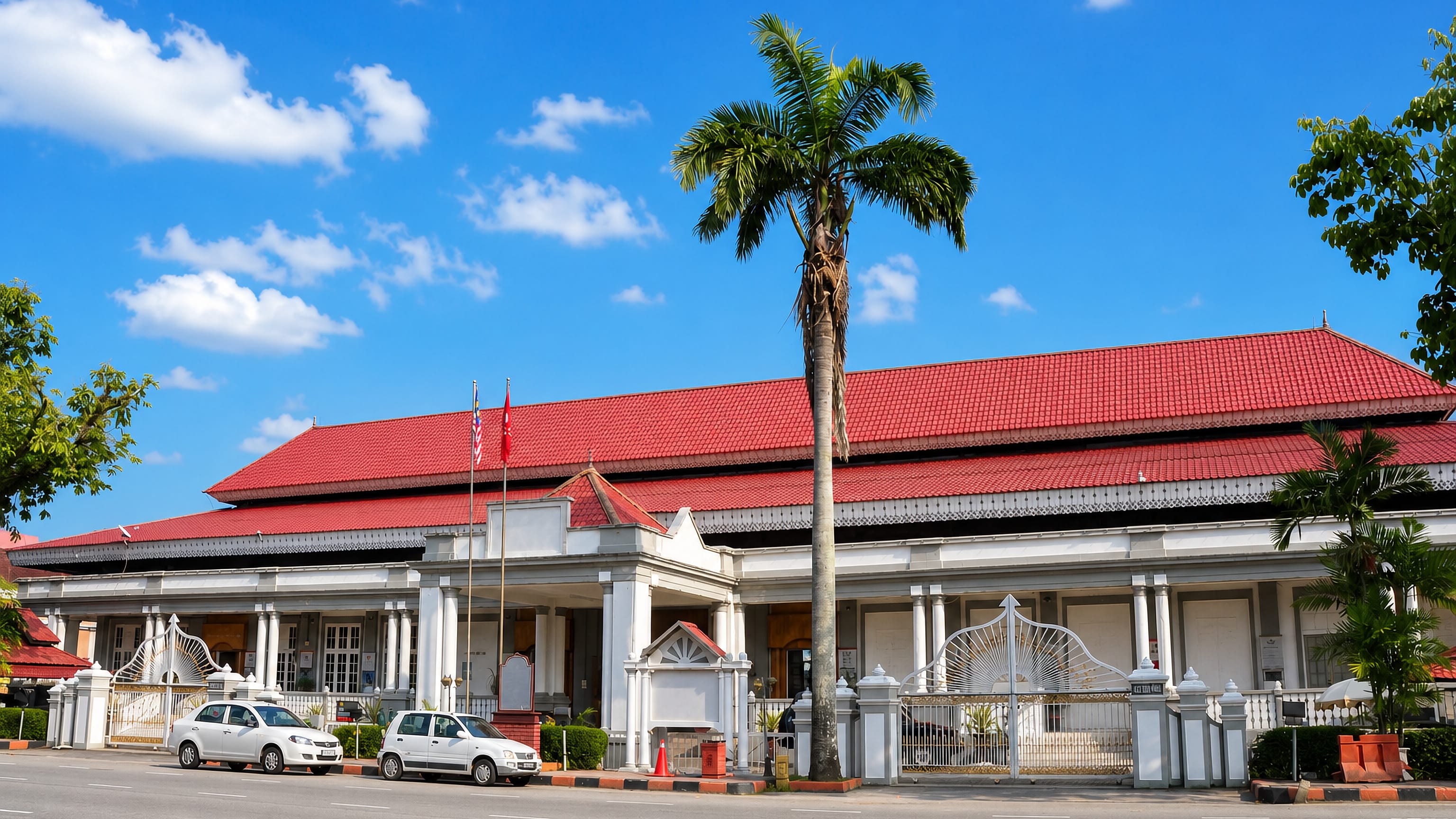 White colonnaded heritage building with a red tiled roof and tall palm on a main street in Kota Bharu.
