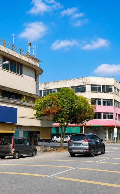 Sunlit shophouse-lined main street in Teluk Intan, Malaysia, with parked cars under a clear blue sky.