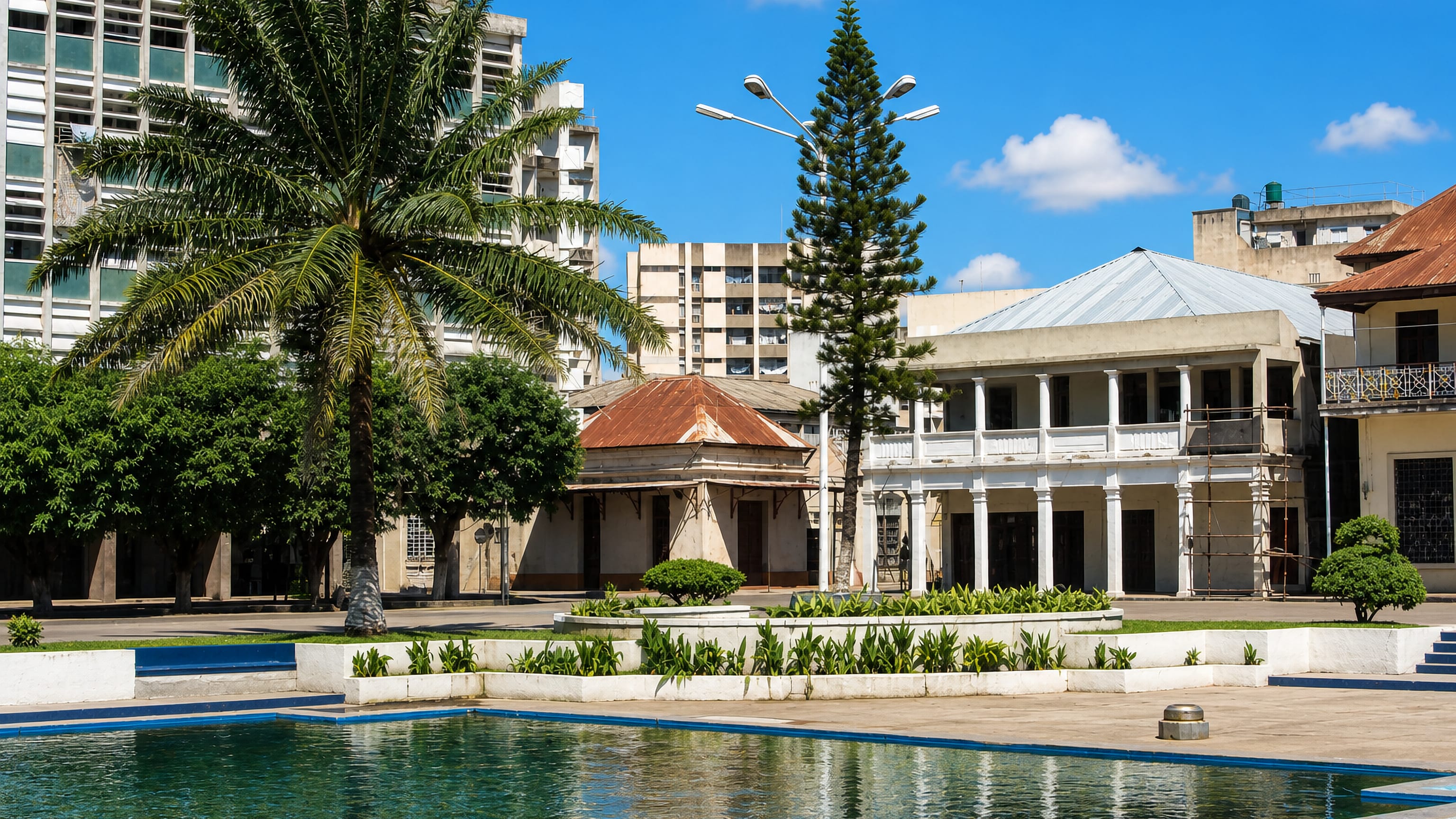 A palm-lined square in Beira with a colonnaded colonial building and modernist blocks behind.