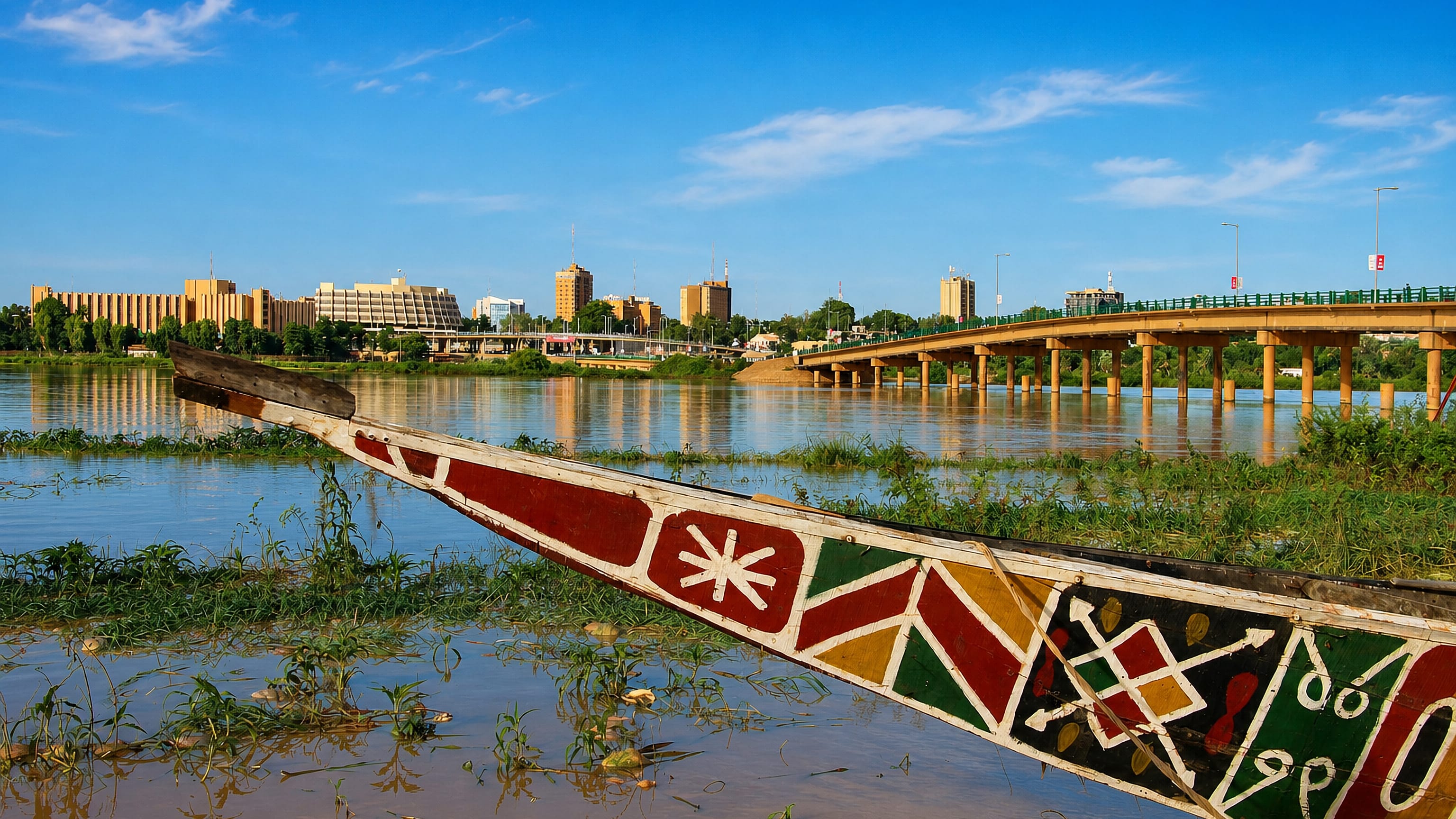 A painted wooden pirogue on the Niger River with the Kennedy Bridge and Niamey's riverfront skyline beyond.