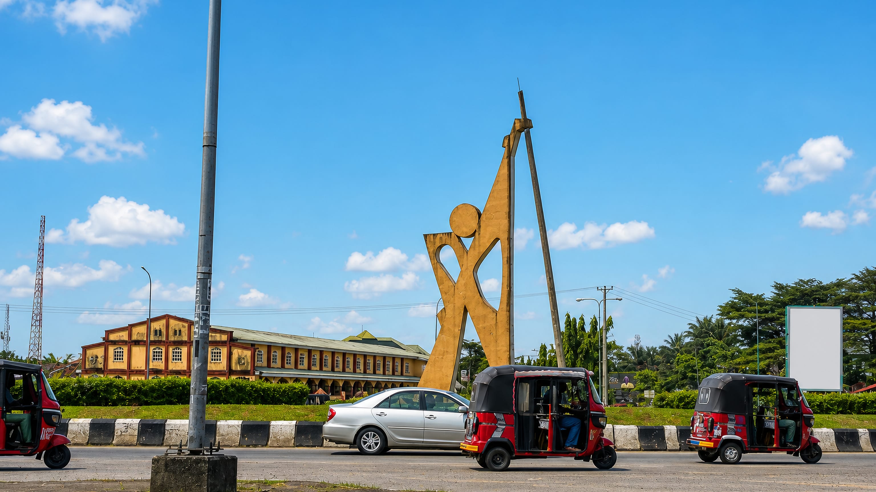 Golden monument on a roundabout with keke tricycles passing a long yellow colonial-style hall in Uyo.