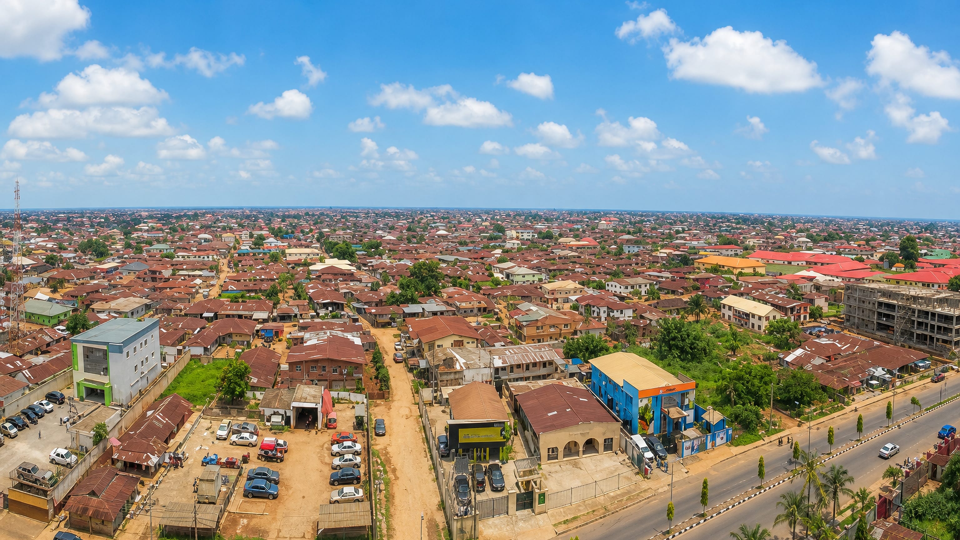 Aerial view over Benin City's rust-red rooftops and tree-lined dual carriageway under a bright blue sky.
