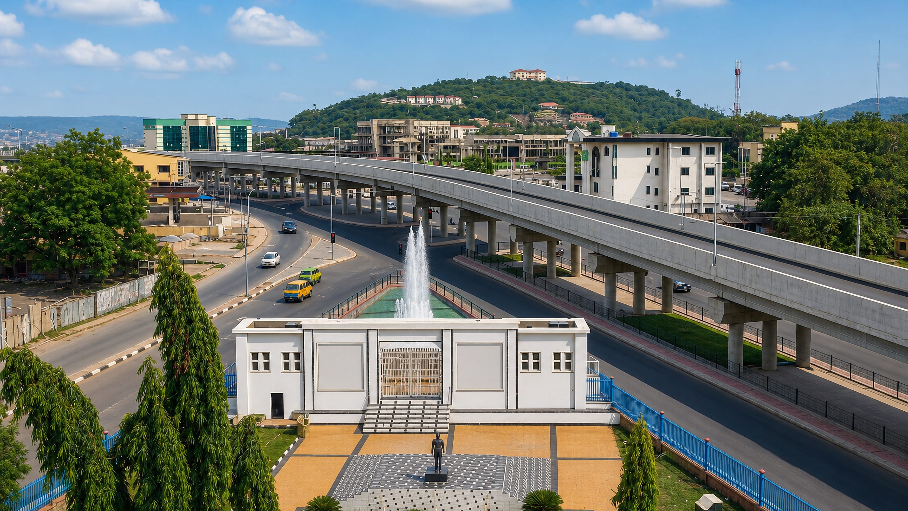 Ado-Ekiti's flyover curving past a fountain and white memorial pavilion, with green hills and hilltop houses beyond.