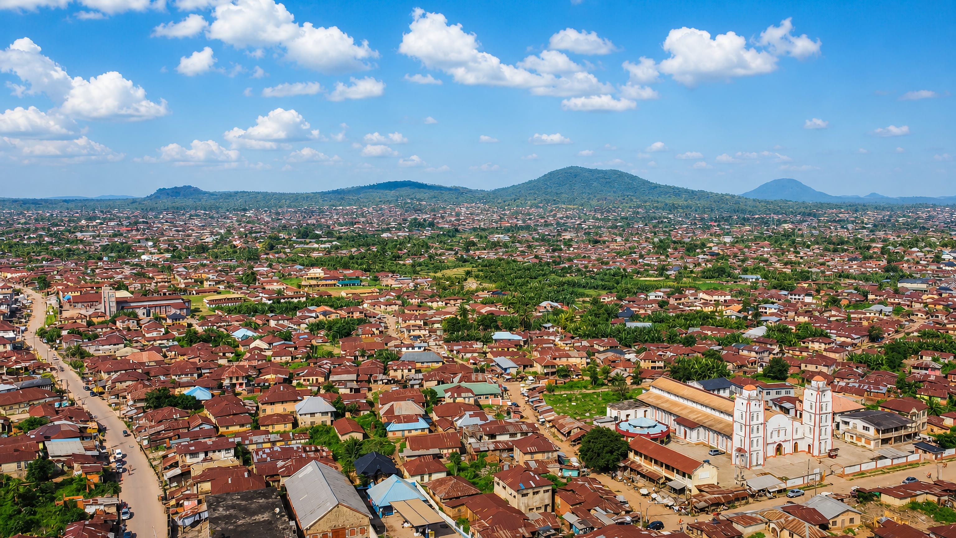 Aerial view over Ile-Ife's rust-red rooftops and a twin-towered church, with wooded hills beyond.