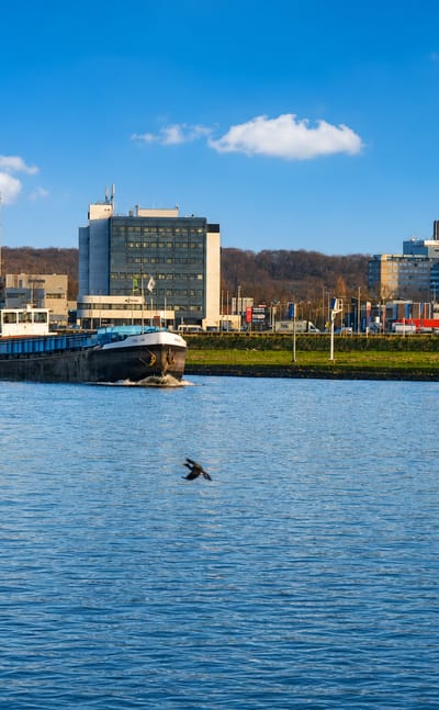 A cargo barge on the Nederrijn river passing Arnhem's riverside offices with wooded hills behind.