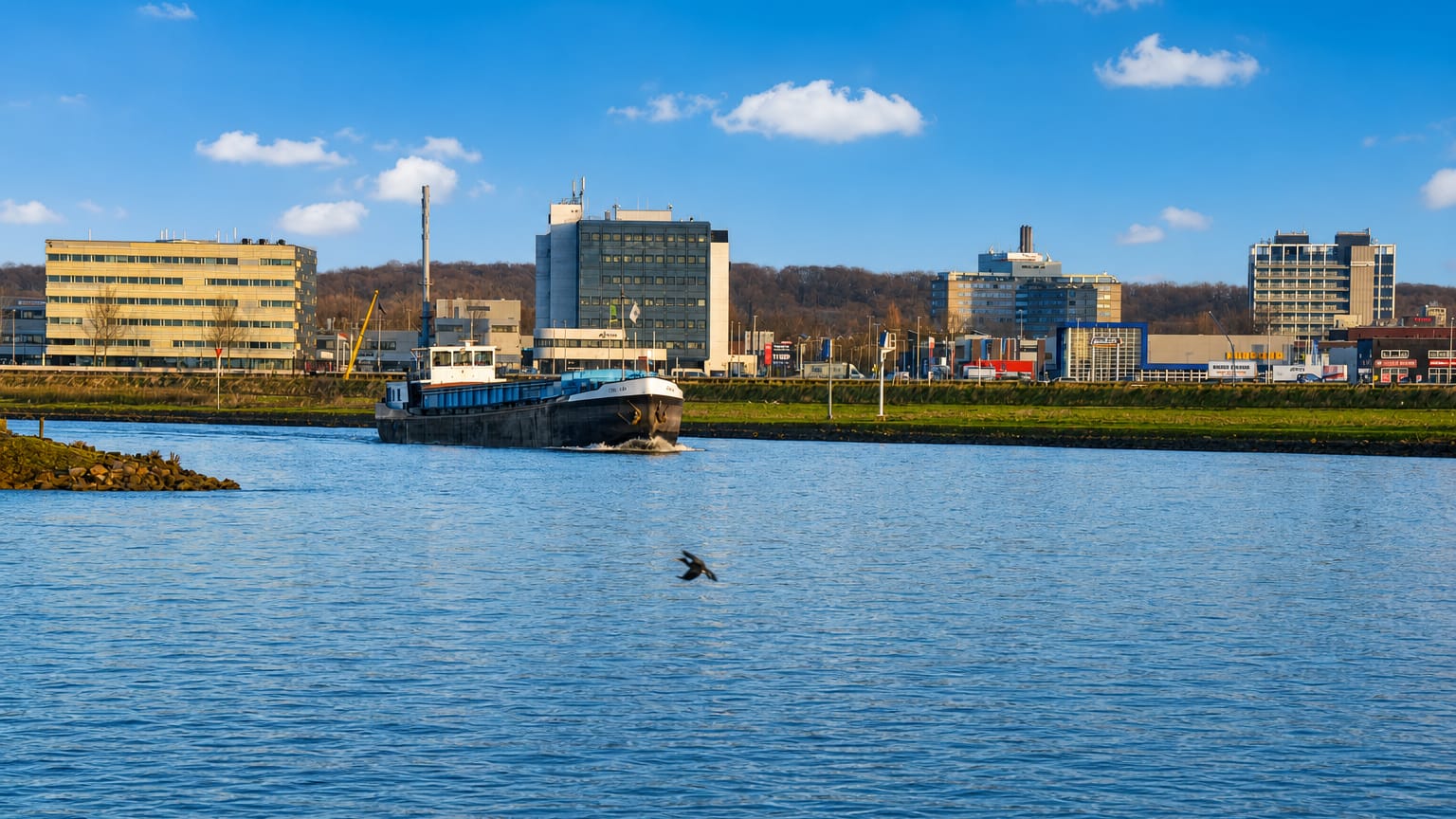 A cargo barge on the Nederrijn river passing Arnhem's riverside offices with wooded hills behind.