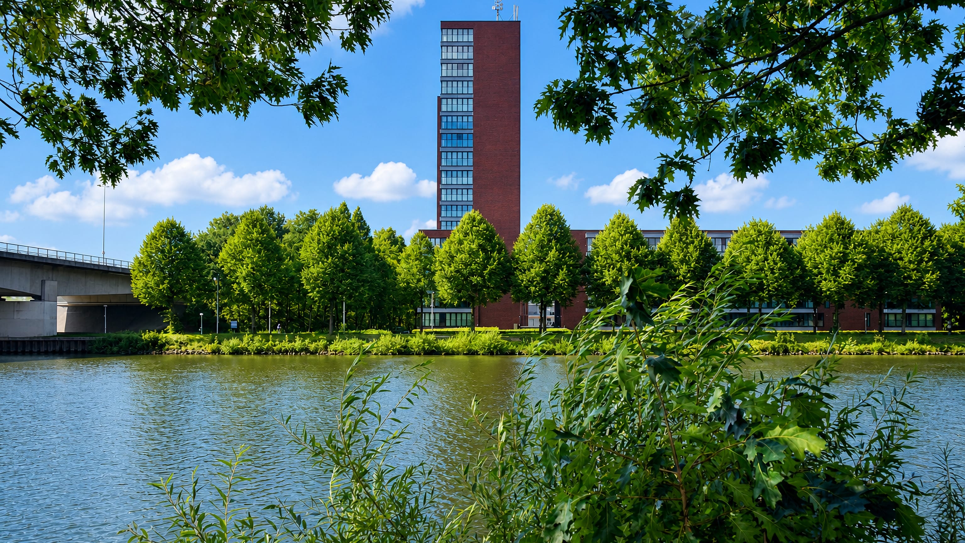 Brick tower block and canal-side bridge seen through summer foliage in Lindenholt, Nijmegen.